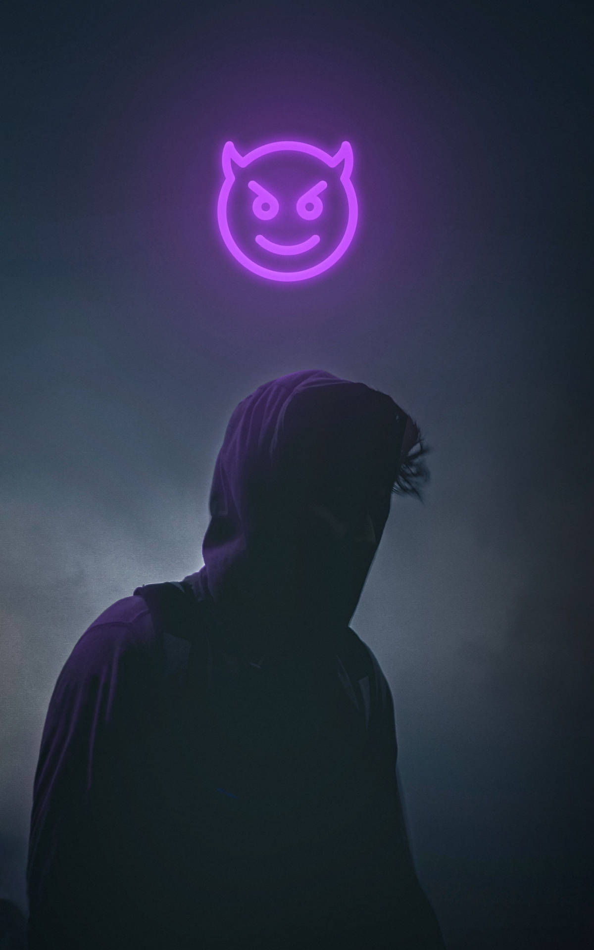 戴兜帽的人在紫色惡魔表情符號霓虹燈牌下的煙霧黑暗中形成剪影。