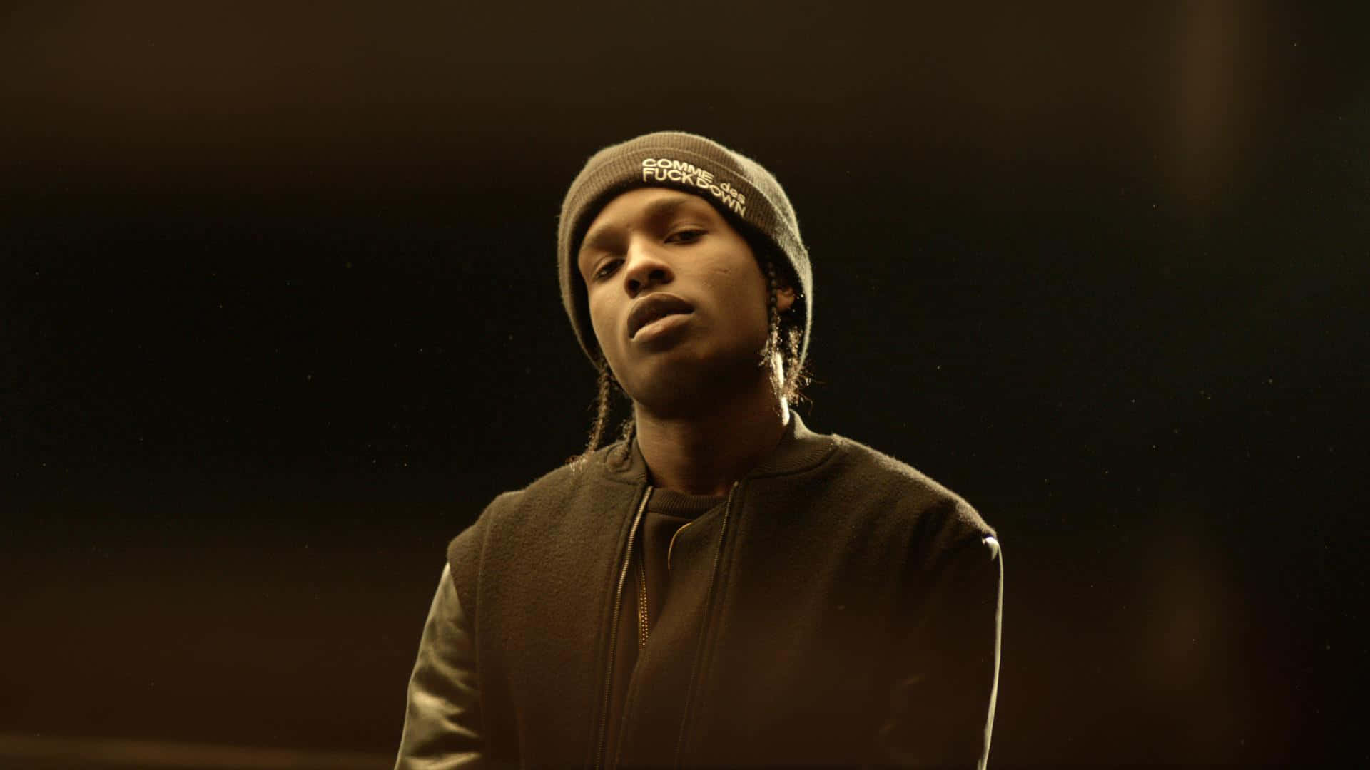 說唱歌手兼時裝設計師 A$AP Rocky 壁紙 — 一個戴冷帽和外套的男人站在昏暗的金色光線中，背景黑暗，有漂浮的粒子。