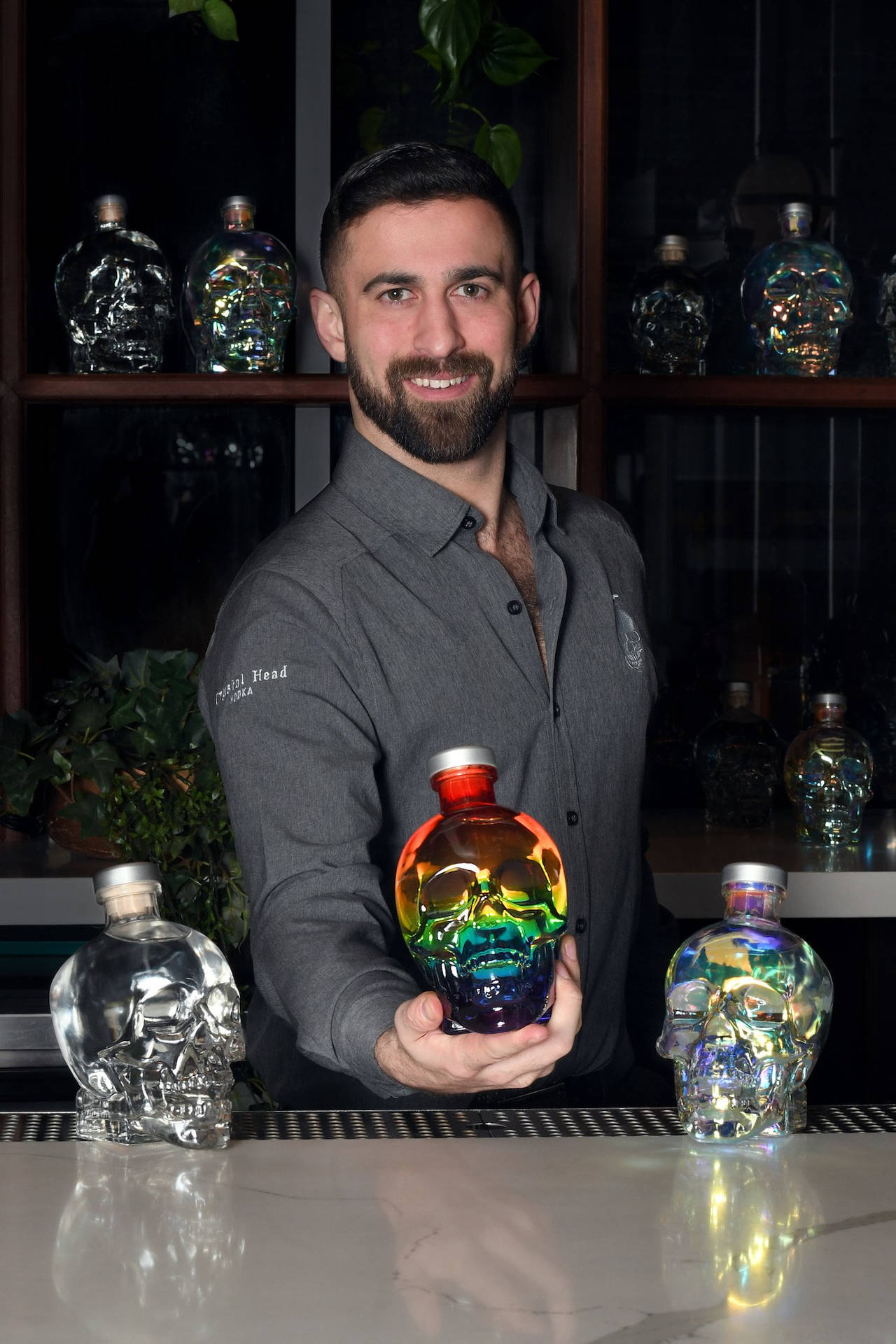 調酒師在吧檯前手持彩虹骷髏瓶，架子上有骷髏利口酒瓶。