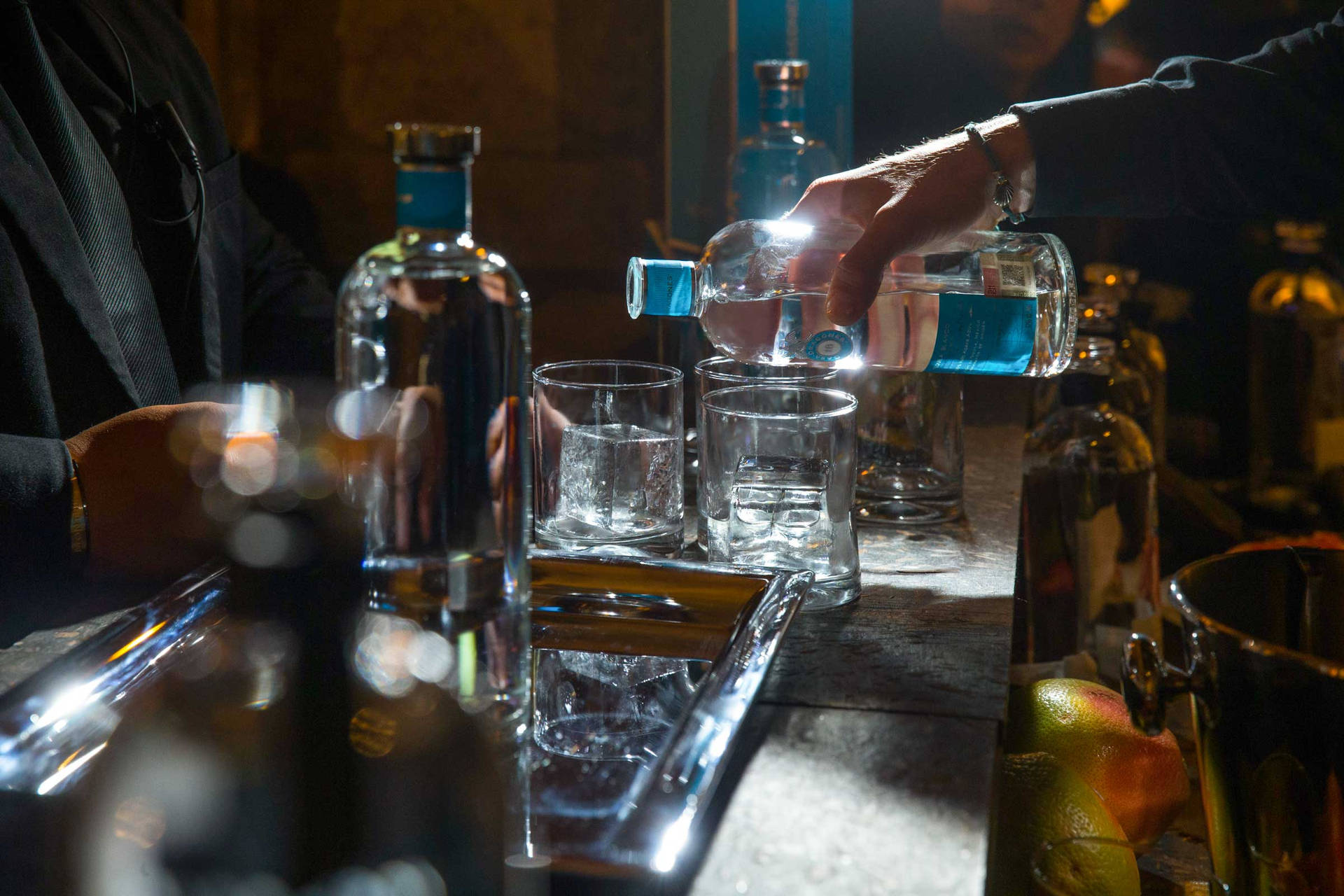 調酒師在燈光昏暗的酒吧中將藍色酒倒入玻璃杯，旁邊有冰塊和柑橘。