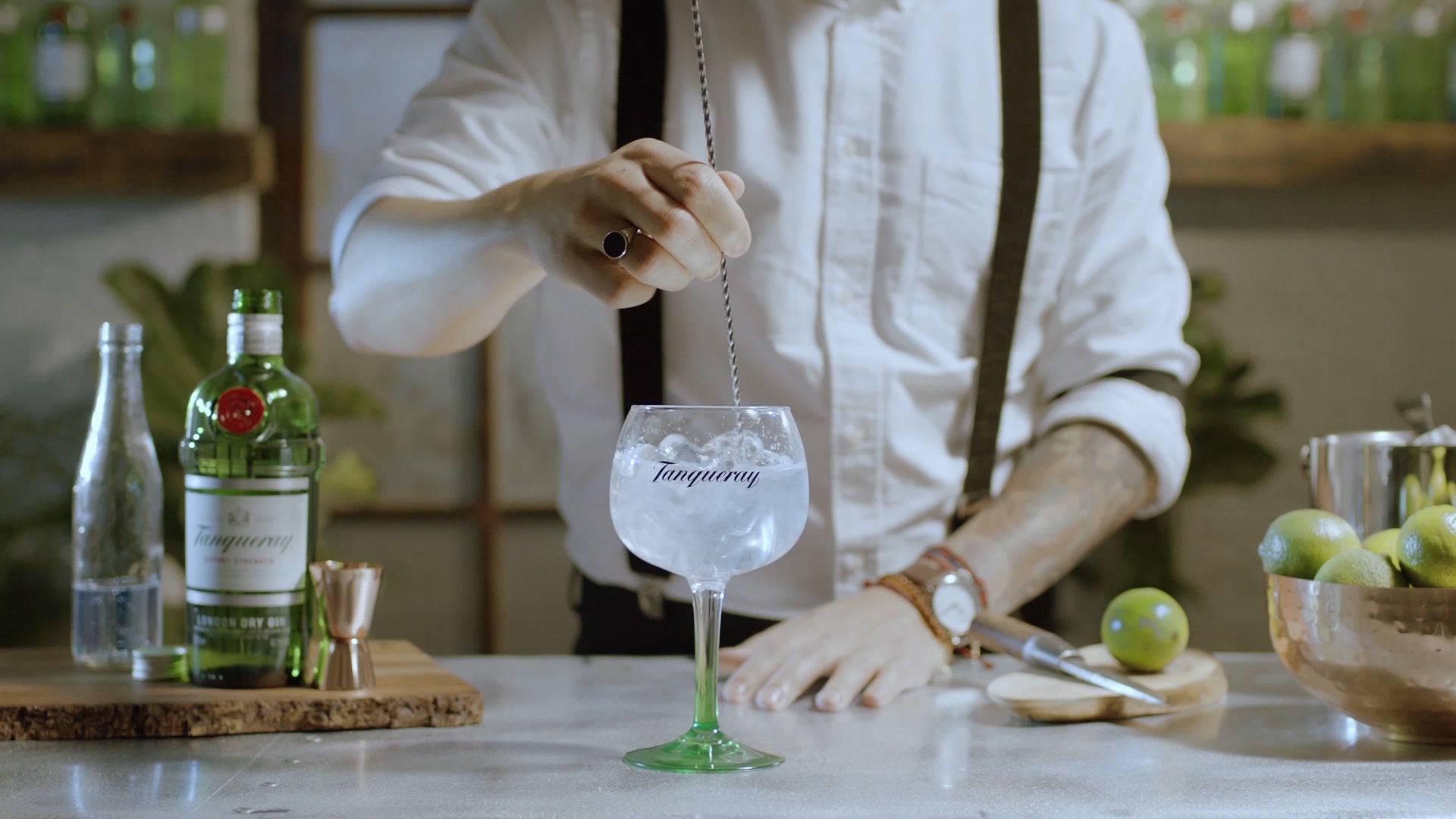 調酒師調製Tanqueray金酒加湯力水 壁紙 — 調酒師在一個鄉村風格的酒吧枱上，用一個品牌玻璃杯攪拌氈酒雞尾酒，周圍有青檸和酒瓶。