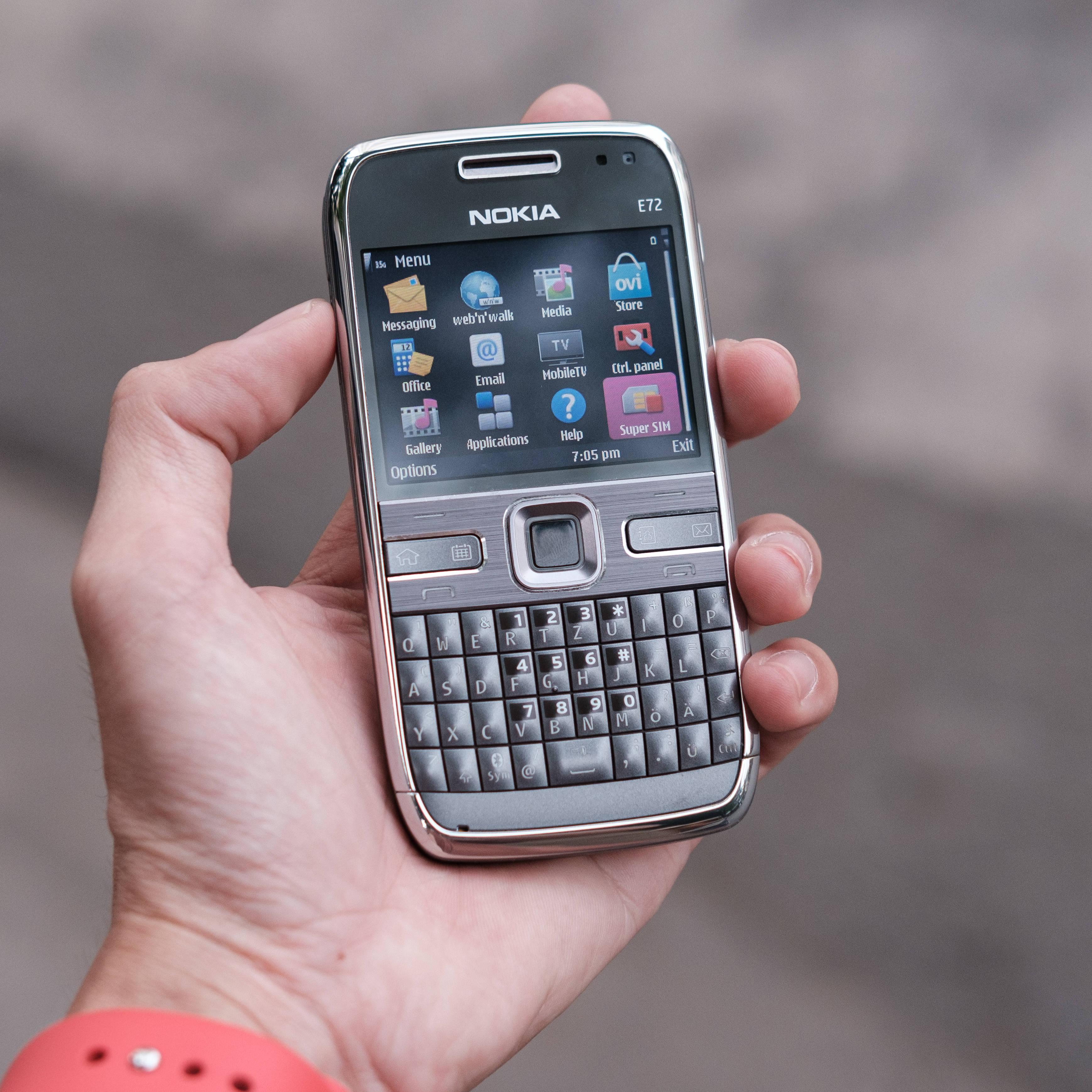 諾基亞 E72 鍵盤手機 壁紙 — 一隻手拿著一部銀色Nokia E72智能手機，帶有實體QWERTY鍵盤和顯示應用程式及時間的活躍屏幕。