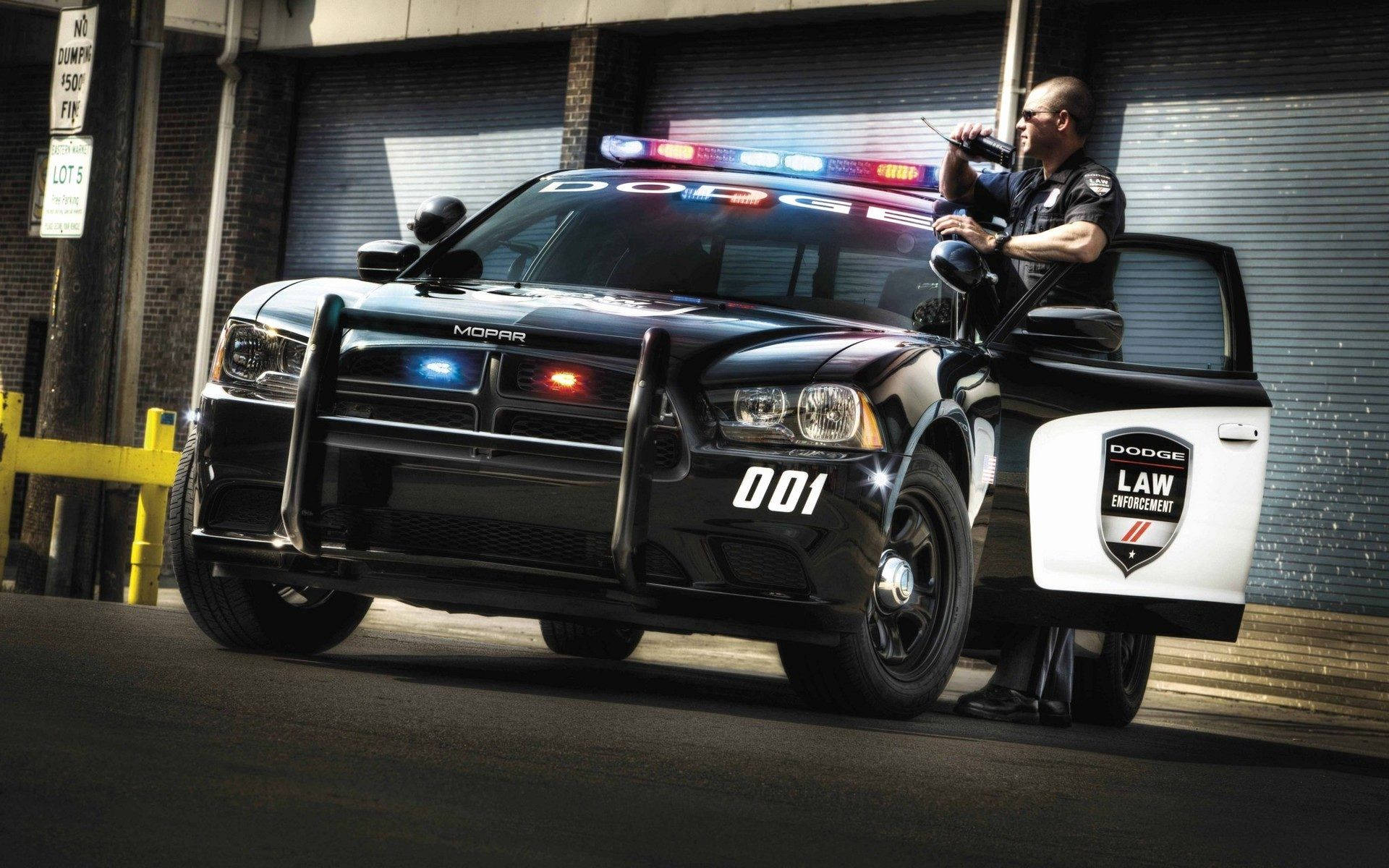 身穿黑色制服的警員從黑色道奇警車的打開車門探出身來，警車在小巷中閃著警燈。
