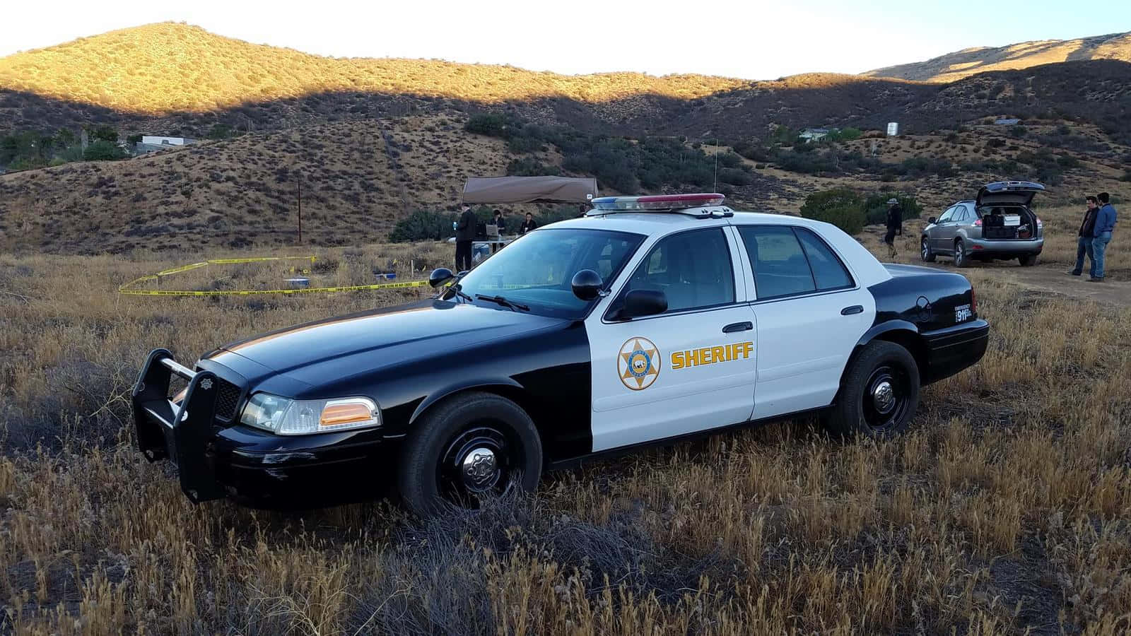 Ford Crown Victoria 桌布 壁紙 — 警長巡邏車停在乾草上，黃色犯罪現場膠帶和調查人員在黃金時段靠近山丘。