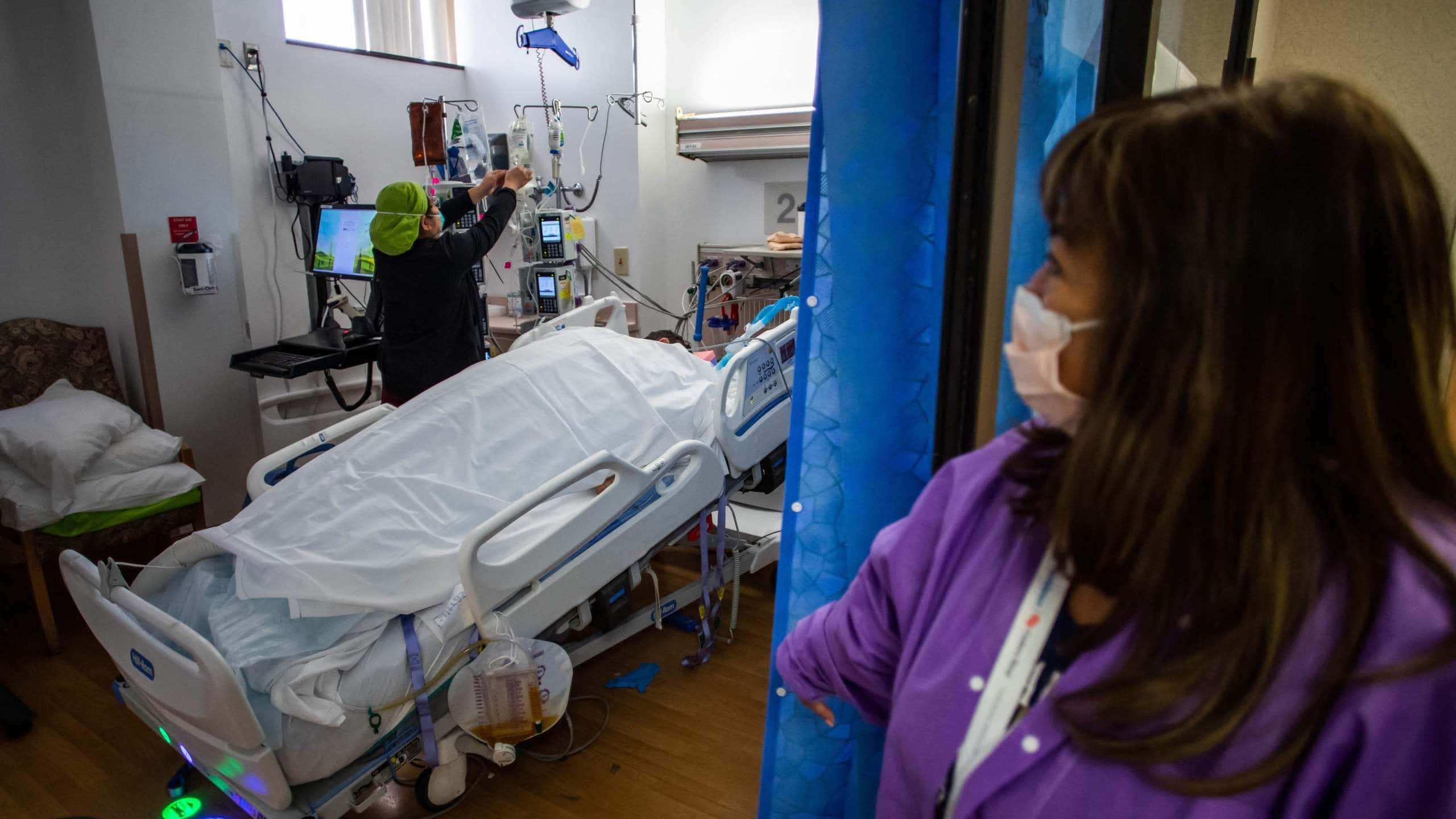 護士凝視病床 壁紙 — 一名戴綠色帽的護士正在醫院病床旁調整醫療設備，另一名戴口罩的工作人員從門口觀察。