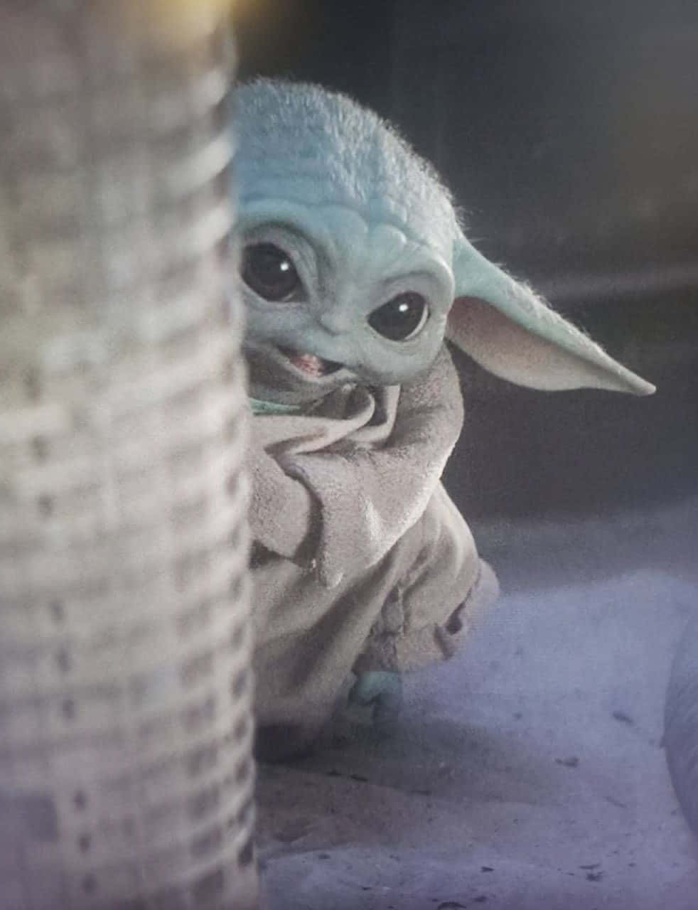 讓Baby Yoda伴你左右 壁紙 — 一個好奇的綠色皮膚嬰兒尤達在昏暗燈光下從編織柱子後面探出頭來。