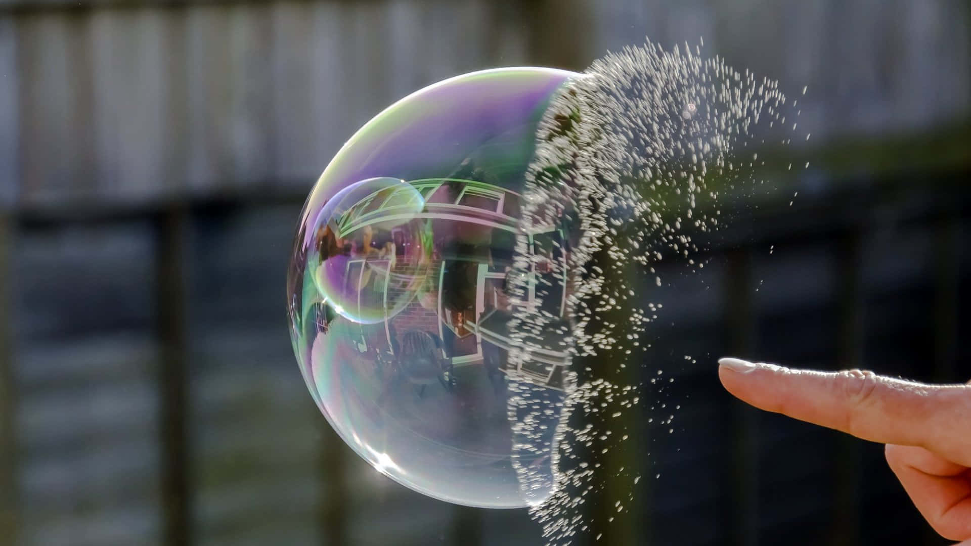 讓你的創意透過 Bubble 來實現 壁紙 — 手指輕輕戳破一個巨大的彩虹色肥皂泡，使其在模糊的木柵欄背景下爆裂成閃閃發光的碎片。