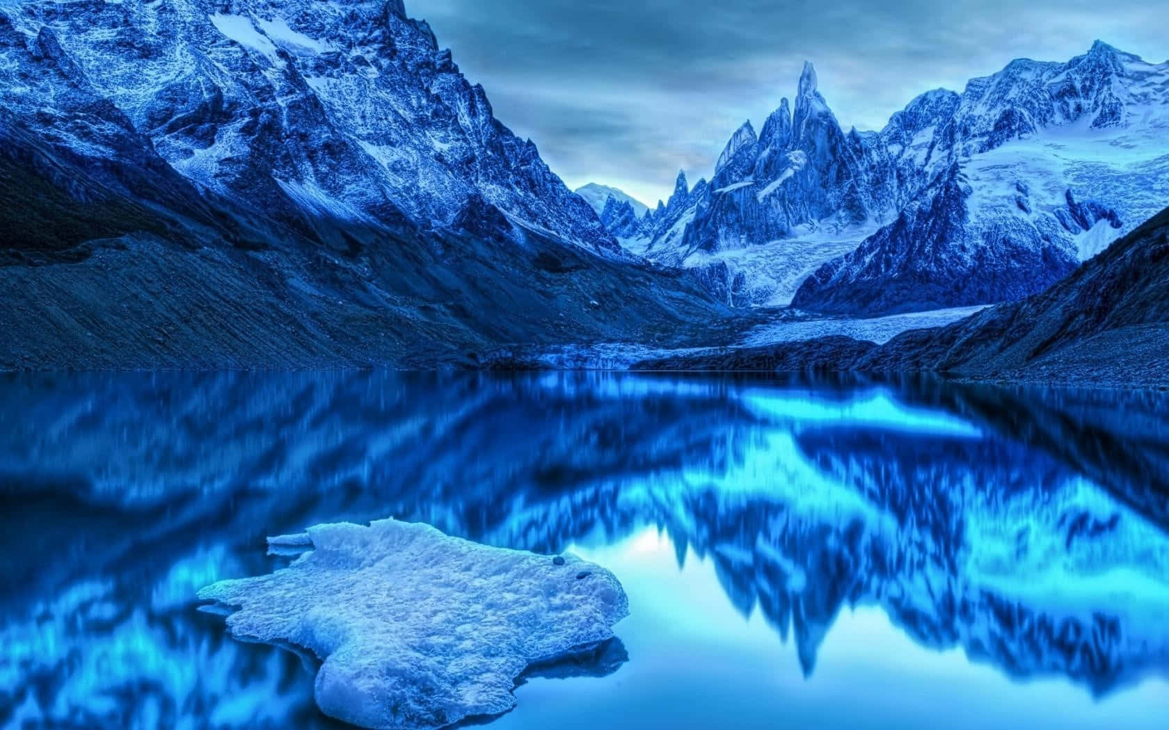 積雪的山峰倒映在平靜的藍色湖水中，附近漂浮著一塊浮冰。