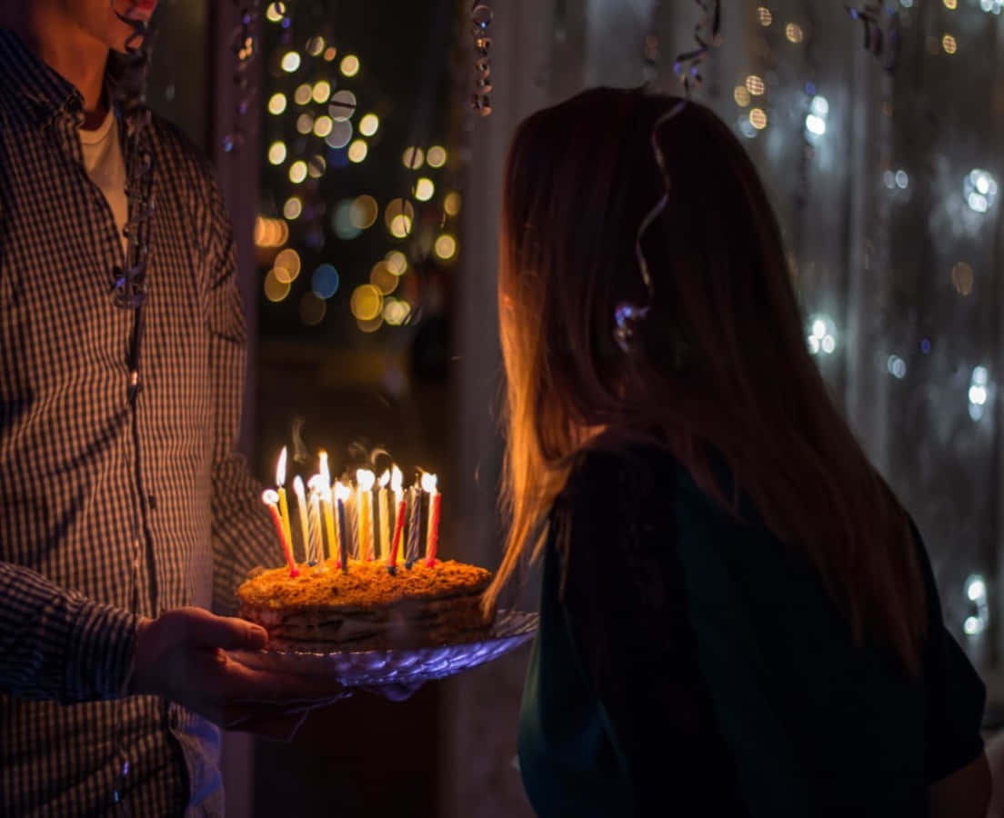 「讓我們慶祝吧！」 壁紙 — 一個人在一個溫暖照明的房間裡，在散景燈串下，向另一個人展示一個點燃蠟燭的生日蛋糕。