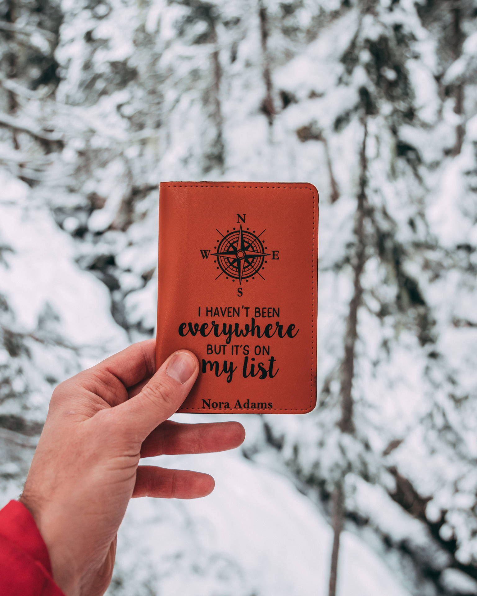 手拿著一個棕色皮革護照套，帶有指南針和旅行名言，在雪覆蓋的森林中。 — 旅行 壁紙