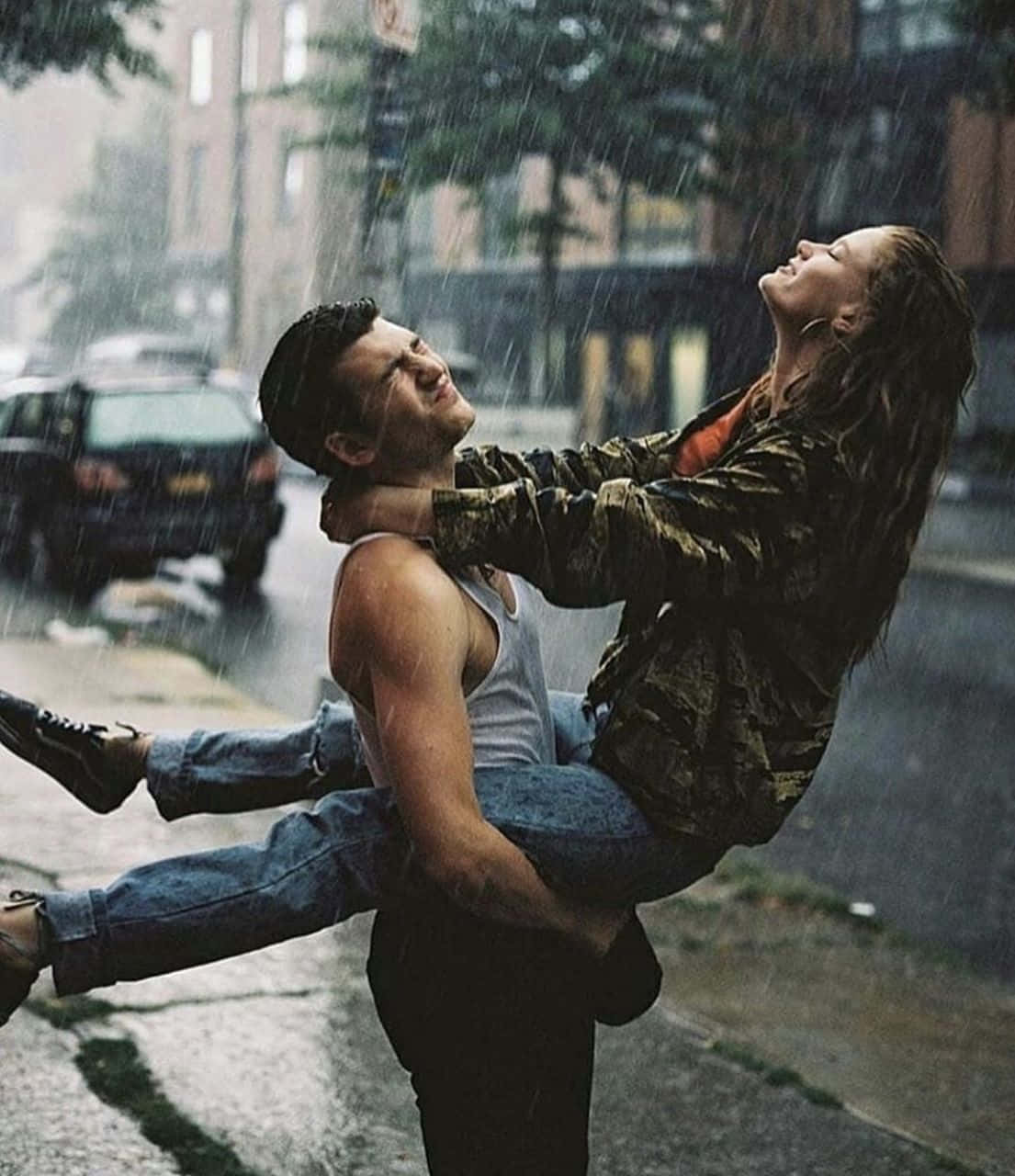 讓雨洗去所有煩惱，提醒我們彼此的愛 壁紙 — 一對情侶在雨中快樂地擁抱，背景是濕漉漉的街道和模糊的汽車。