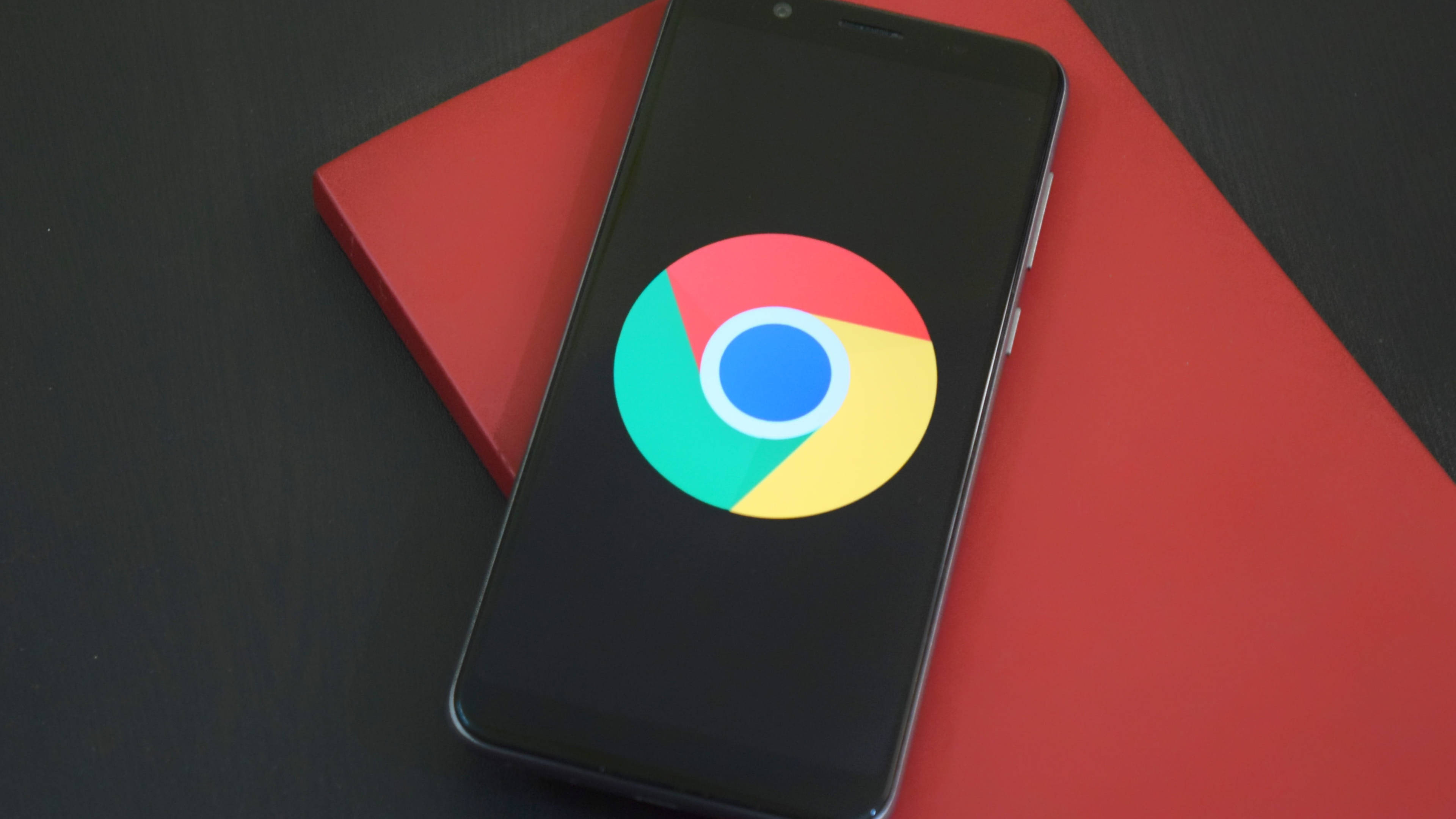 谷歌 Chrome 手機版 壁紙 — 一部顯示Google Chrome標誌的智能手機放在深色表面上的鮮紅色書本上。