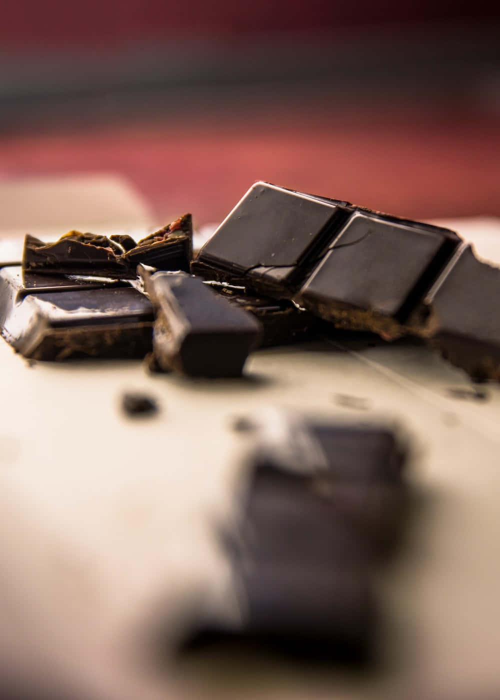 Dark Chocolate 桌布 壁紙 — 豐富的黑巧克力碎片，具有光澤質感和破碎邊緣，放置在柔軟的米色表面上，背景為模糊的紅色。