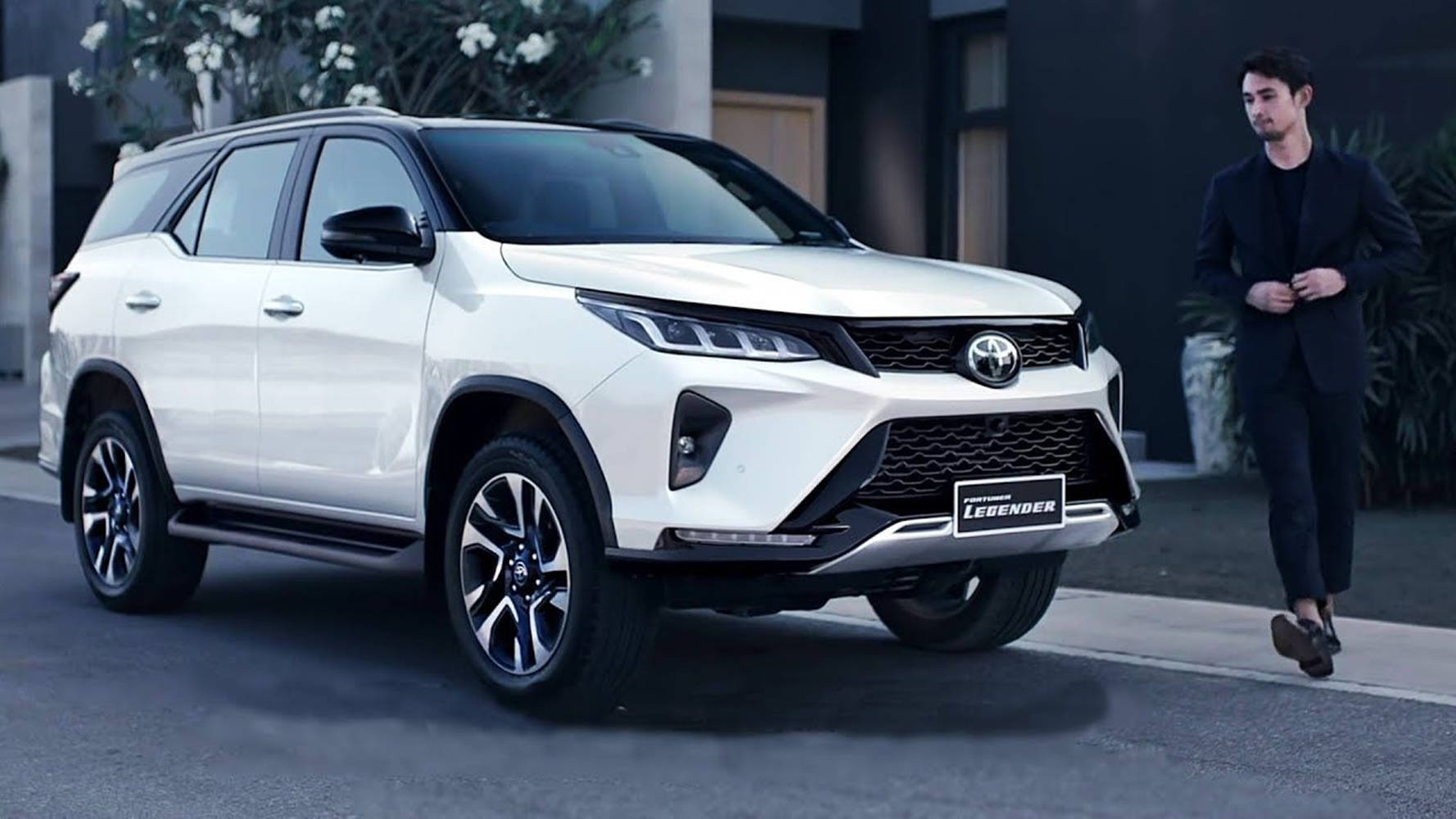 一架白色豐田 Fortuner Legend SUV 停泊在現代街道上，附近有一位身穿黑色西裝的男子走過。