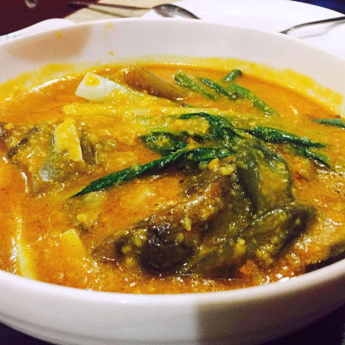 豐盛的菲律賓 Kare-Kare 燉菜 壁紙 — 一碗冒著熱氣的橙色咖喱，裡面有魚塊和綠色香草，在溫暖的燈光下閃閃發光。