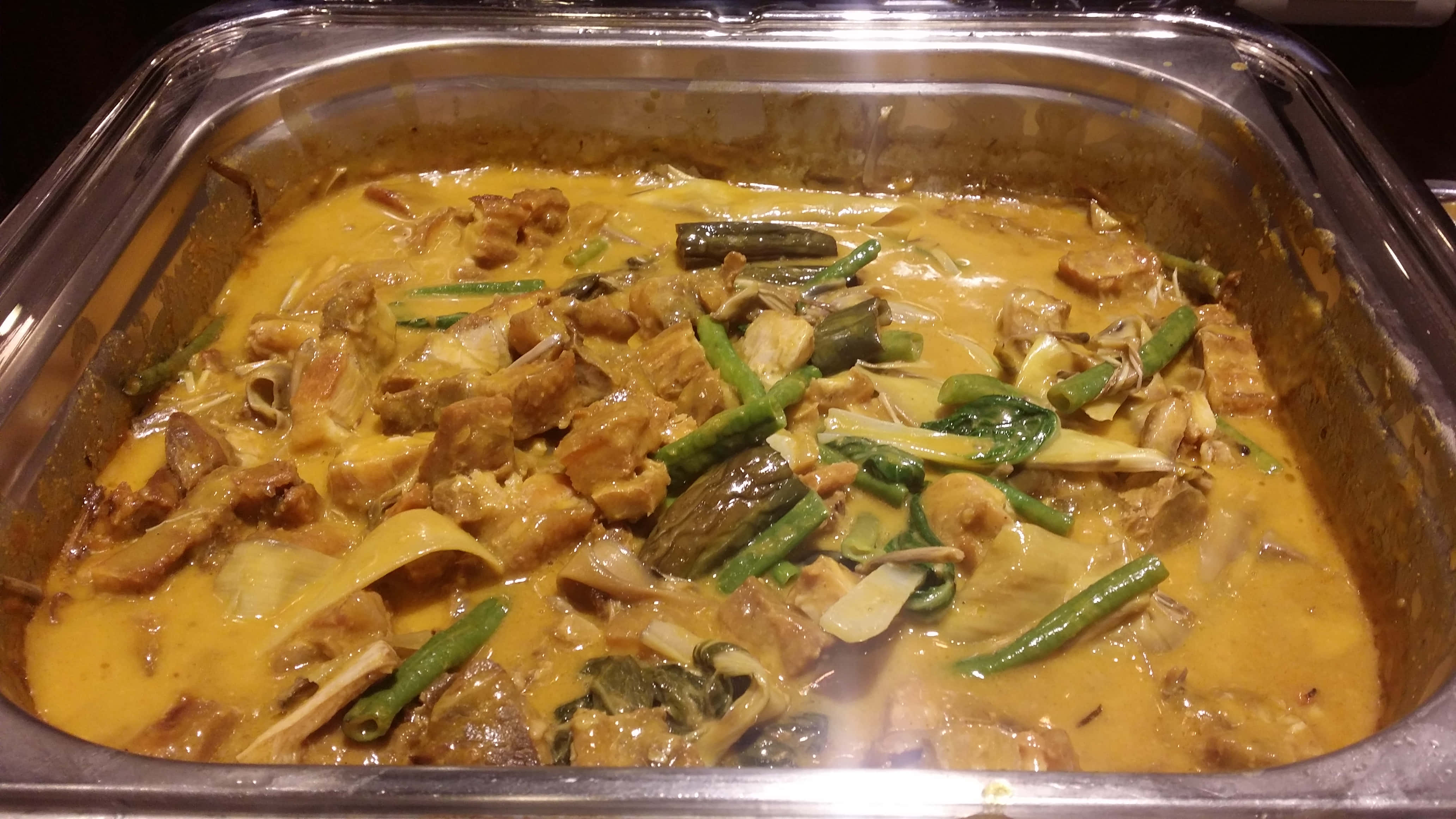 豐盛的菲律賓燉菜 Kare-Kare 壁紙 — 豐富的金色咖哩，裡面有肉塊、青豆和綠葉蔬菜，在不鏽鋼自助餐托盤中冒著蒸氣。