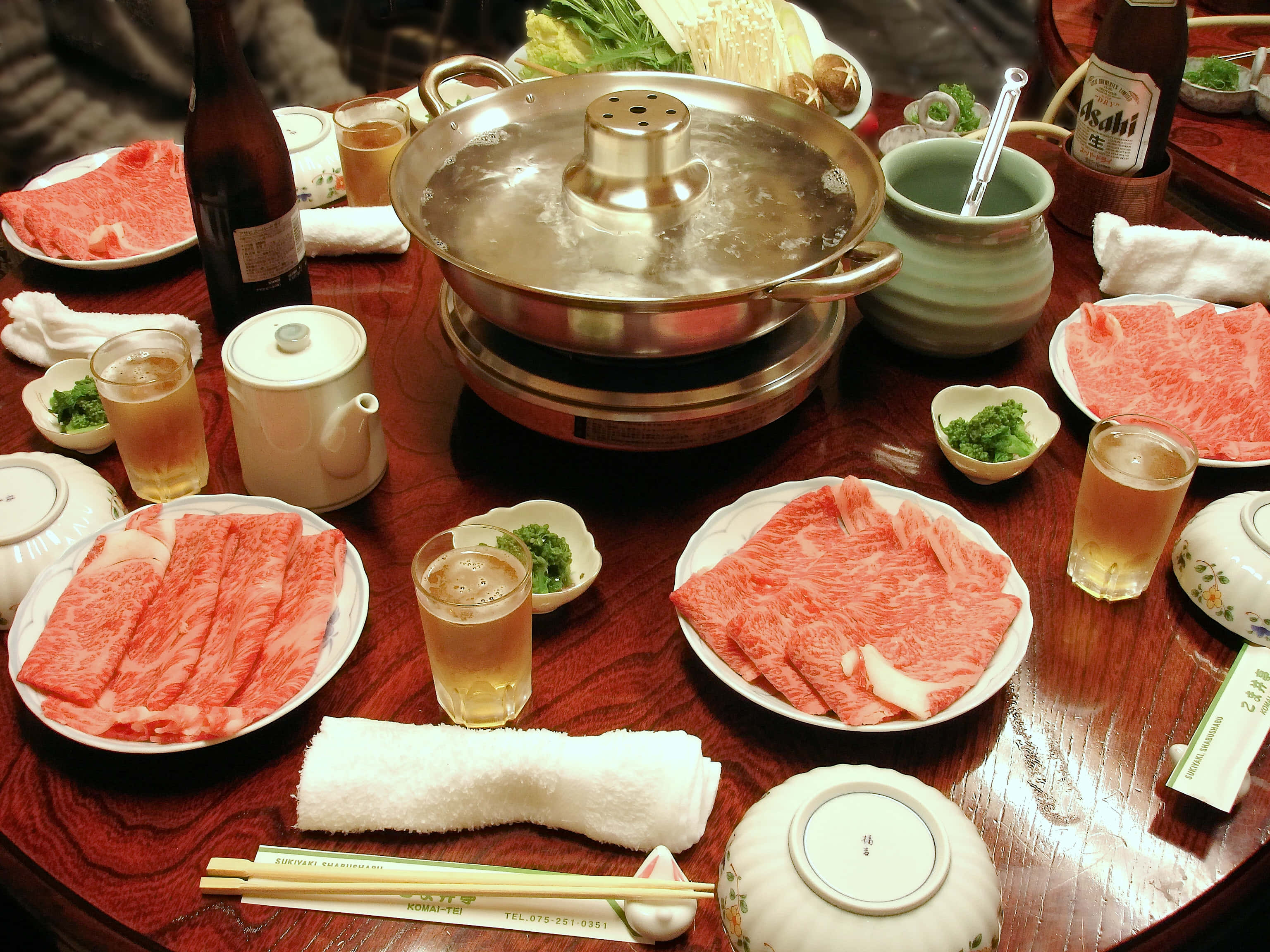 豐盛肉類涮涮鍋 壁紙 — 一個冒煙的熱鍋，周圍放著幾碟生牛肉片和飲料，放在木桌上。