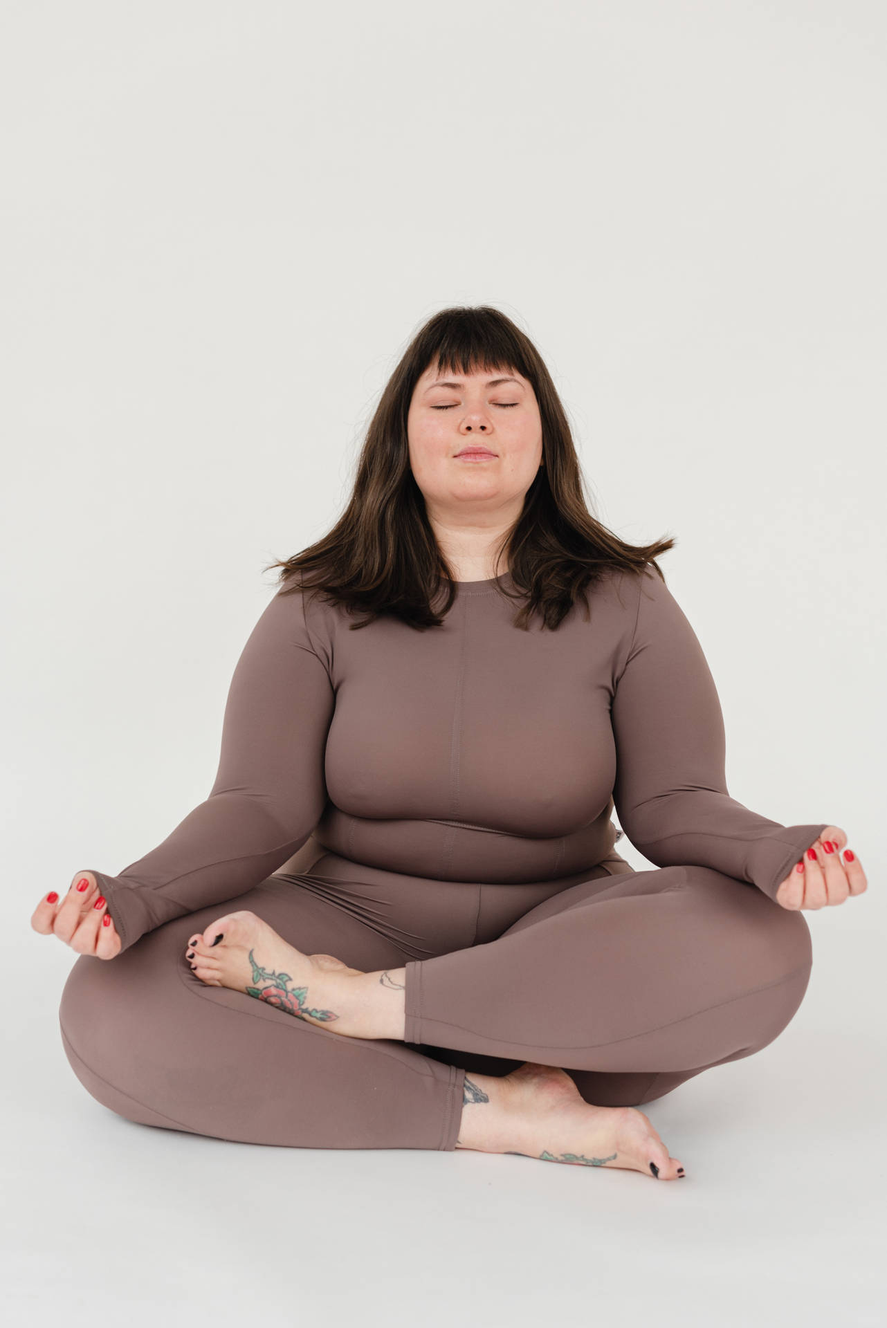 女性身穿棕色服裝盤腿冥想，帶有紋身和紅色指甲。