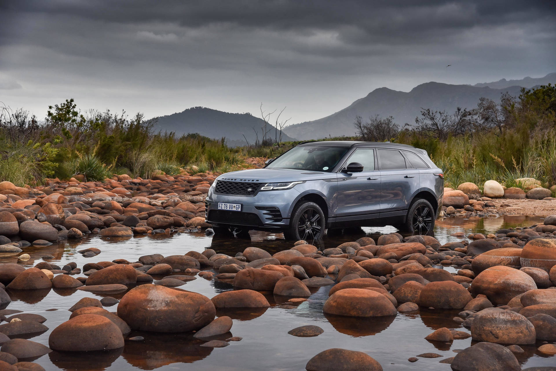 銀色Range Rover Sport在淺水岩石溪流中行駛，背景有山脈。