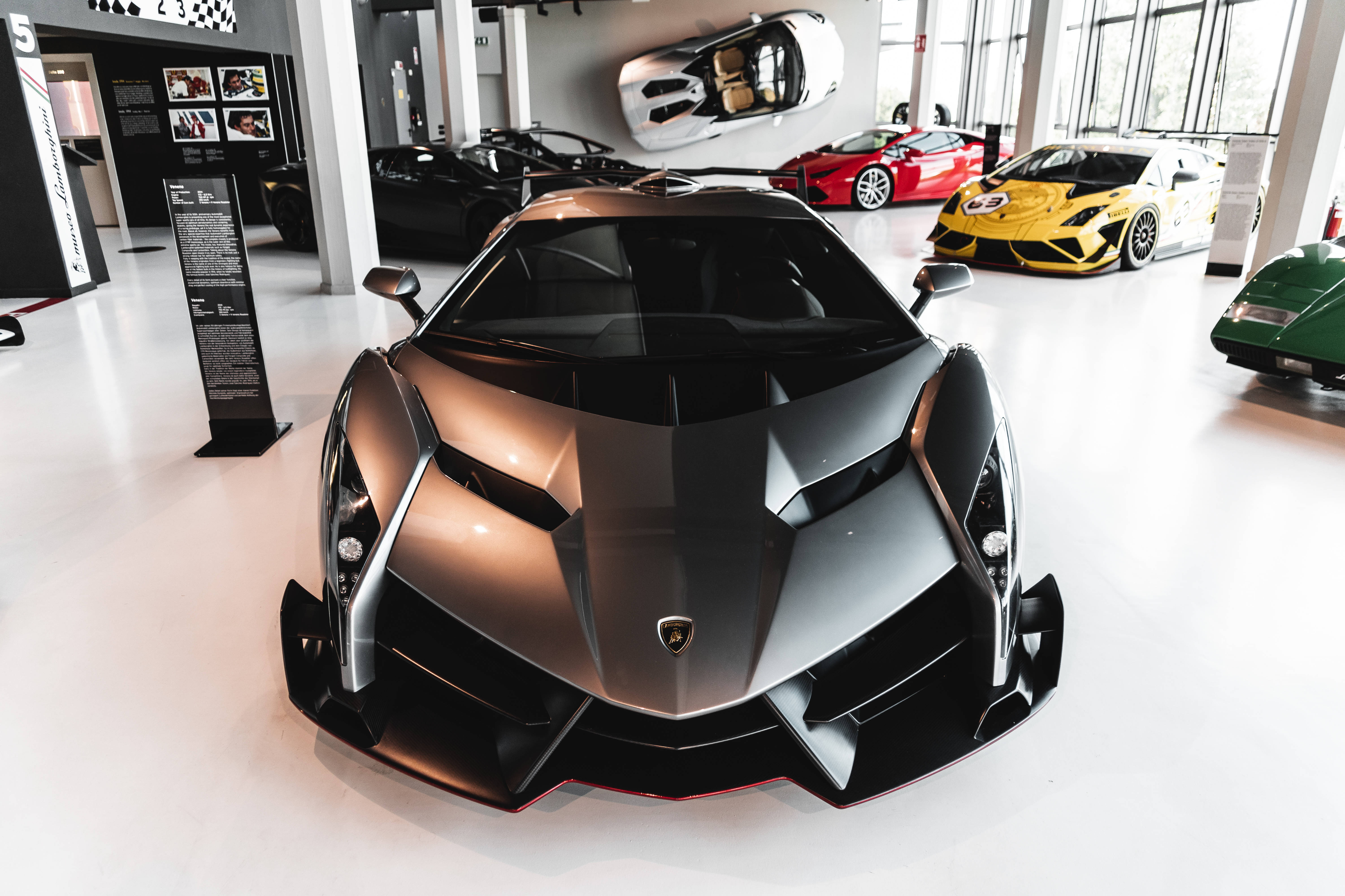 「豪華Veneno V12在汽車博物館的驚豔展示」 壁紙 — 一輛線條流暢的黑色林寶堅尼 Veneno，帶有銳利角度和紅色點綴，在明亮的現代汽車展廳中與其他豪華跑車一同佔據主導地位。