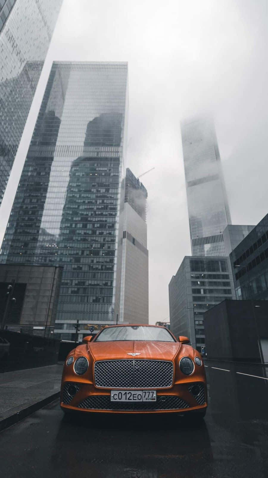 橙色 Bentley 停泊在多雨城市的高大玻璃建築之間。