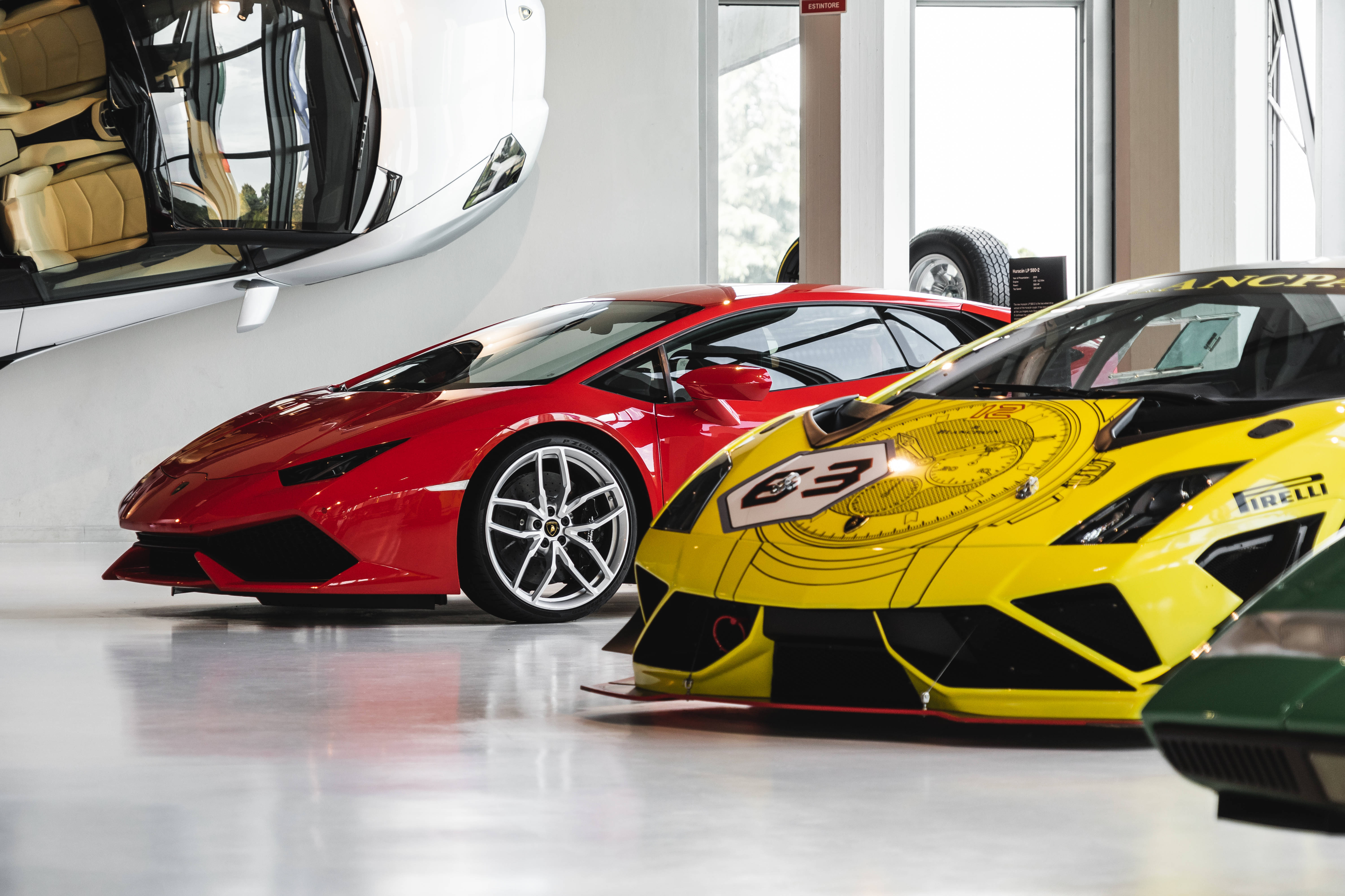 豪華汽車博物館Lamborghini車隊 壁紙 — 兩輛流線型林寶堅尼超級跑車，一輛紅色街車和一輛黃色賽車，在明亮的燈光下於潔白的車庫中閃閃發光。