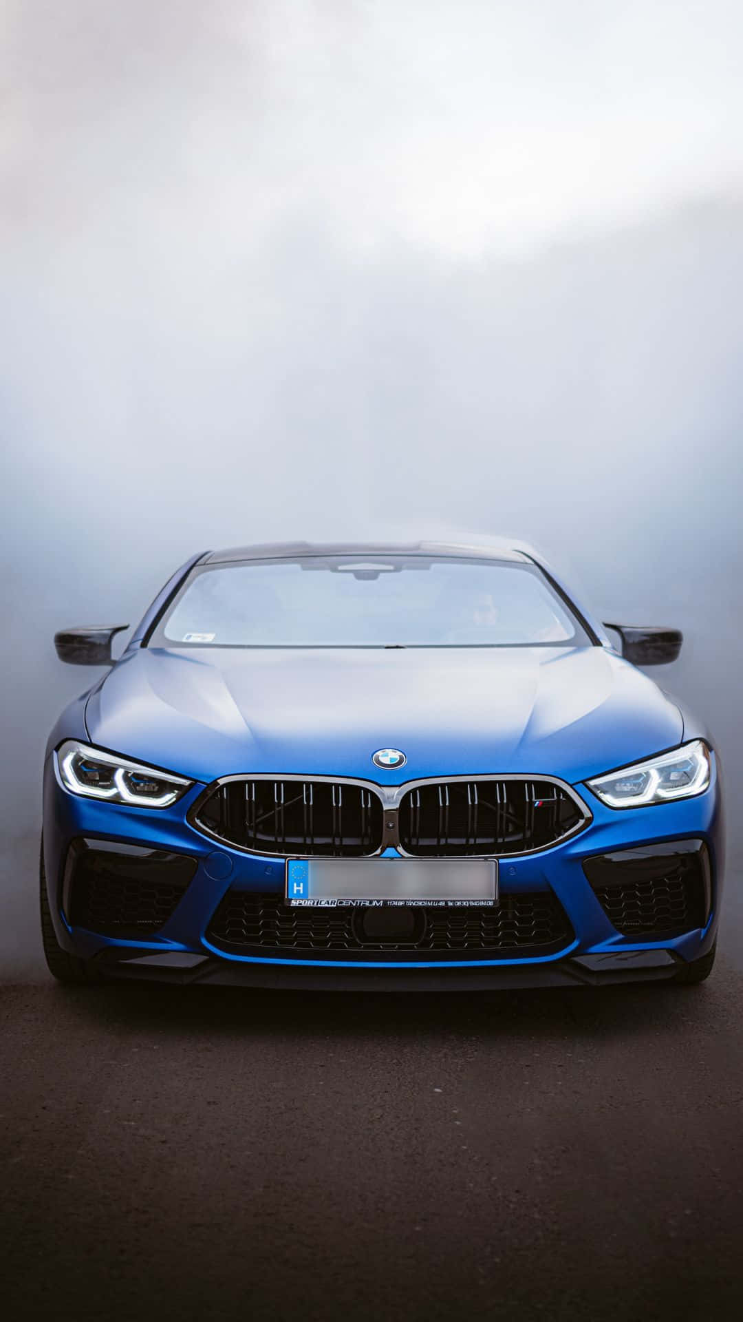 豪華汽車的未來 壁紙 — 一輛引人注目的藍色BMW M8 Coupe在柔和光線下閃閃發光，其侵略性的前格柵和鋒利的頭燈主導了迷霧、極簡的工作室環境。