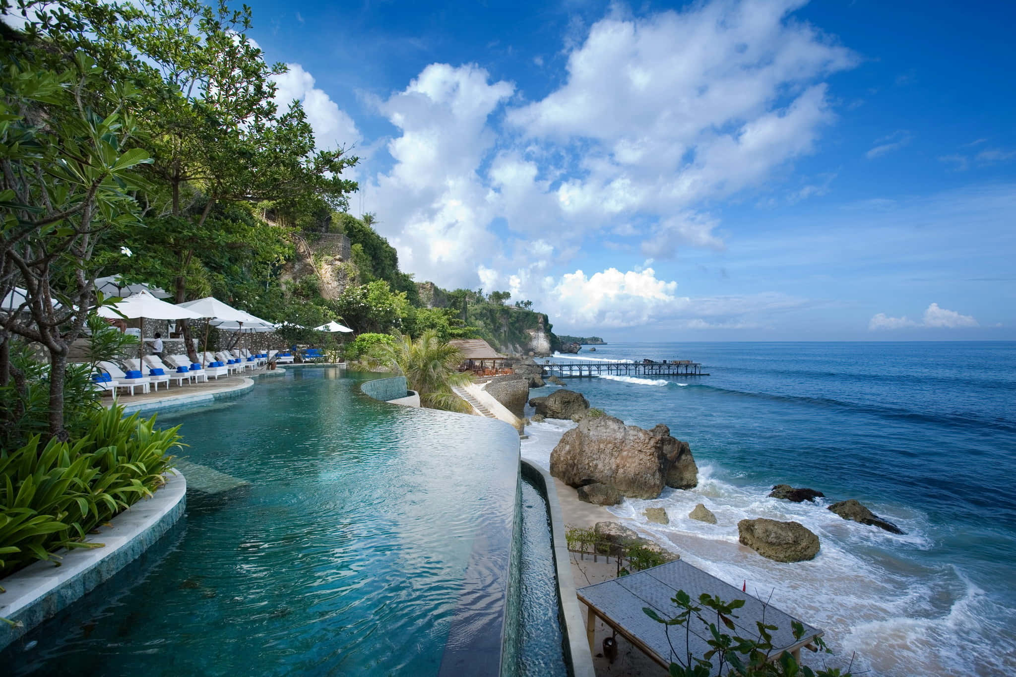 Bali 桌布 壁紙 — 豪華的無邊際泳池與碧綠色的海洋無縫融合，背景是鬱鬱蔥蔥的綠色懸崖和明亮的藍天。