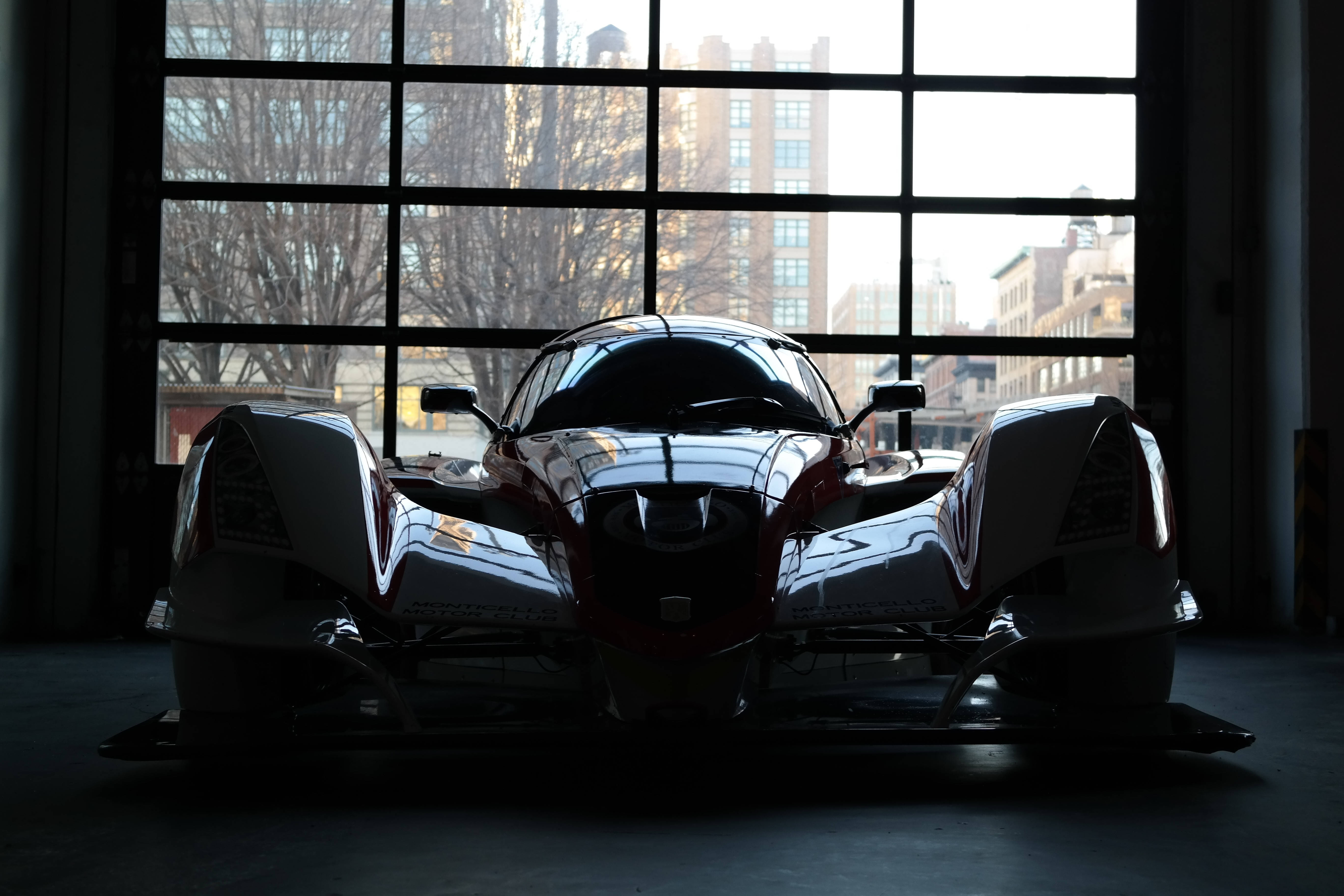豪華賽車 壁紙 — 一架時尚的未來賽車在倉庫內呈現剪影，其反光表面捕捉到大窗戶的光線，窗外顯示光禿的樹木和城市建築。