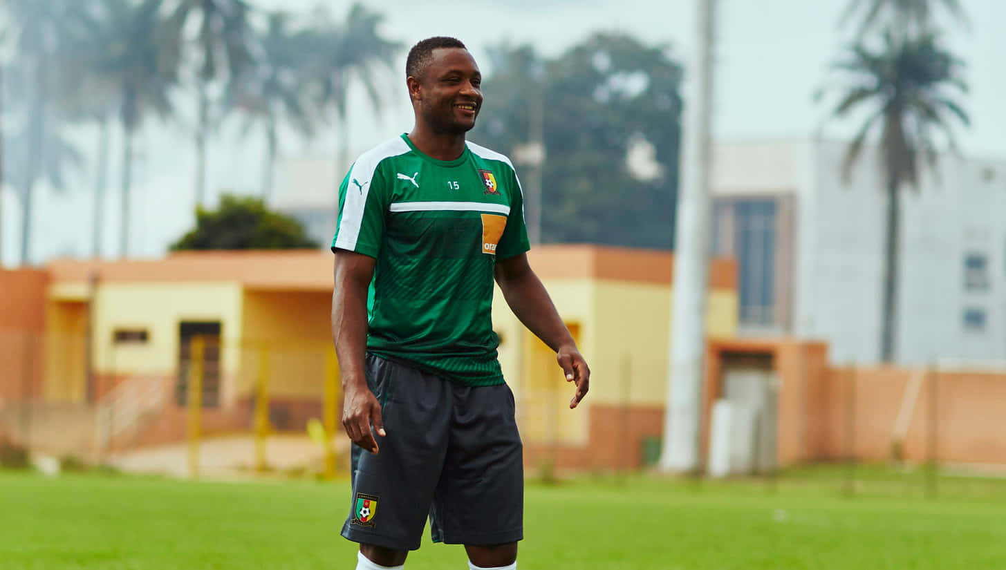 豹隊簽下前 Roger Milla 壁紙 — 一位微笑的足球運動員身穿綠色球衣，站在草地上，背景有棕櫚樹和建築物，柔和。