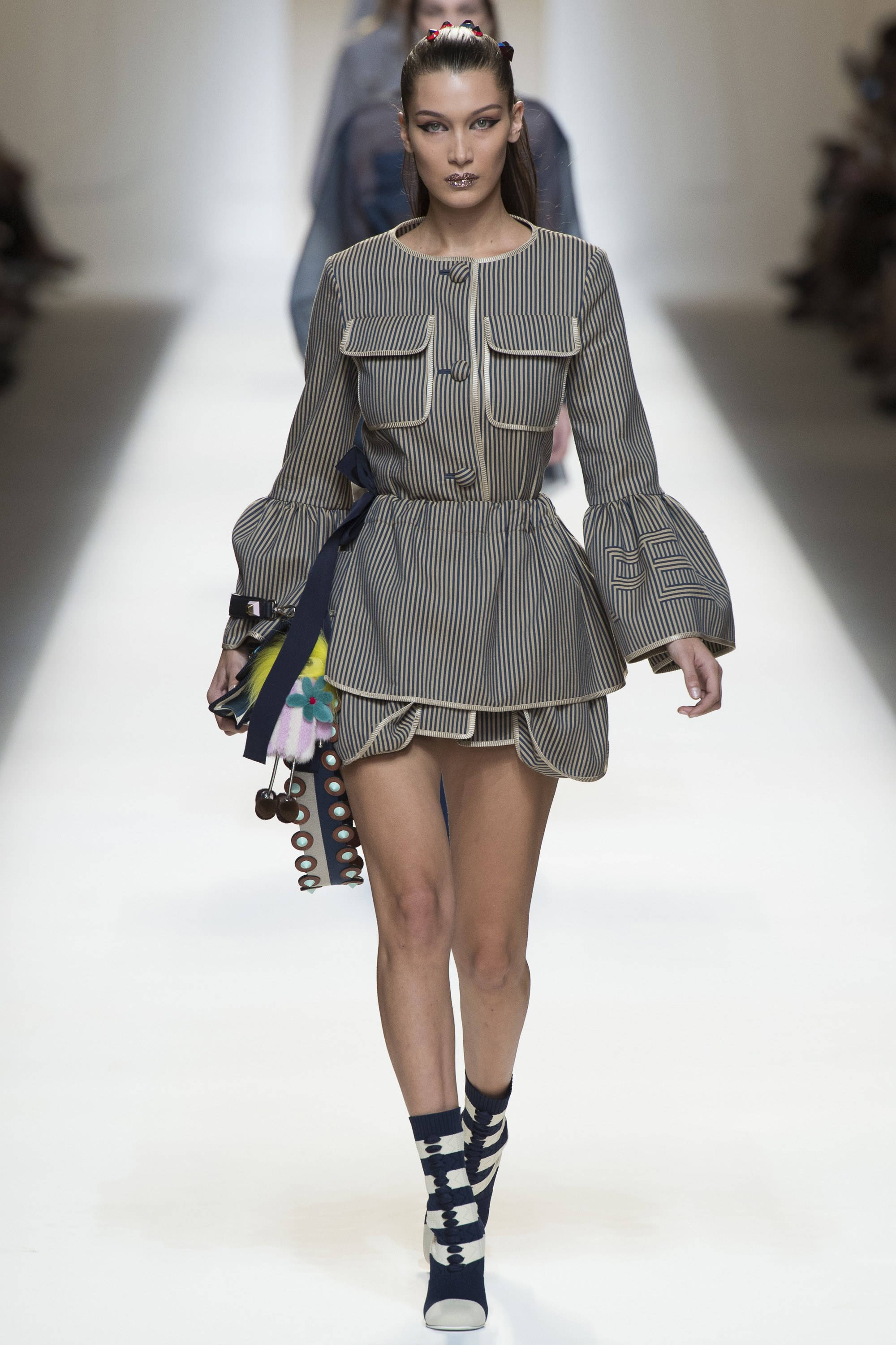 貝拉·哈迪德穿 Fendi 連衣裙 壁紙 — 模特兒自信地走在明亮的白色天橋上，身穿大膽的條紋荷葉邊上衣，搭配喇叭袖和圖案靴子。