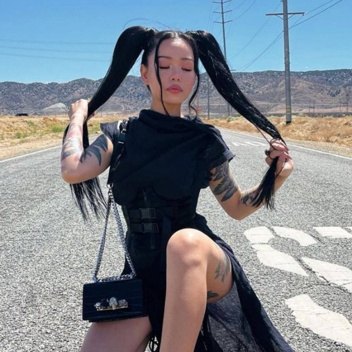 貝拉·波奇TikTok名人 壁紙 — 一位留著黑色長辮子、有紋身的女性坐在沙漠公路上，在清澈的藍天下抓著頭髮。