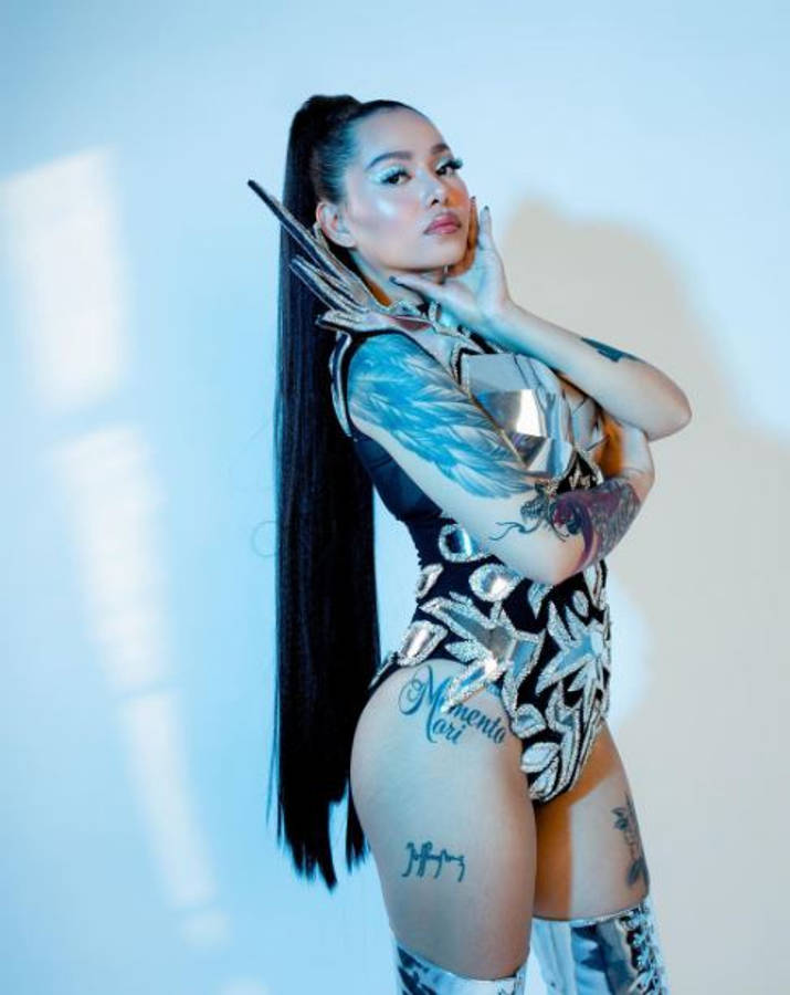 貝拉·波奇服裝造型 壁紙 — 一位擁有長黑髮和紋身的女性，在冷藍色燈光下，穿著金屬緊身衣，自信地擺姿勢，背景是純白牆壁。