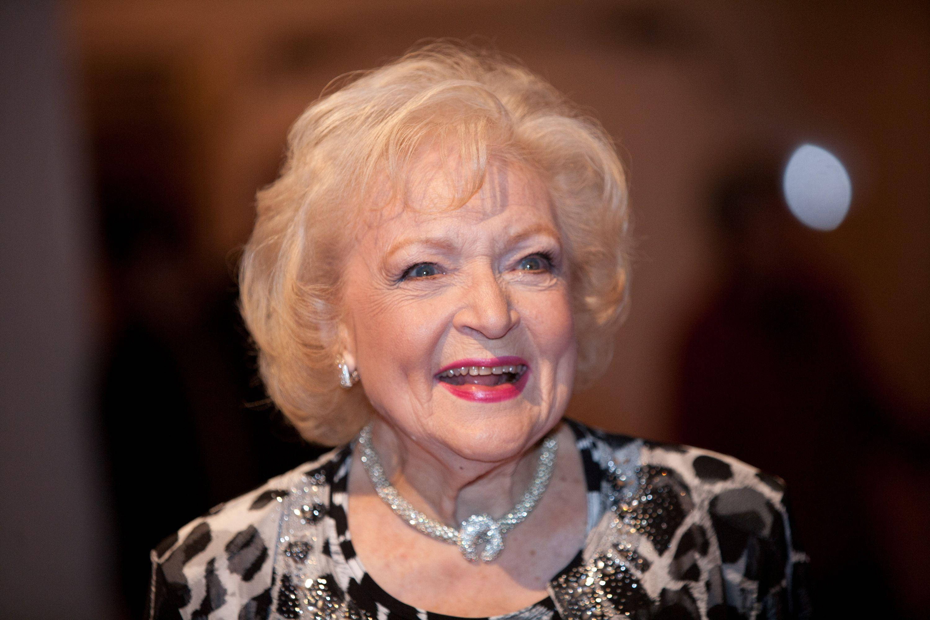 貝蒂·懷特參加美國喜劇獎典禮 壁紙 — 一位滿面紅光的銀髮女士，金髮紅唇，穿著閃爍的黑白套裝，溫暖地微笑。