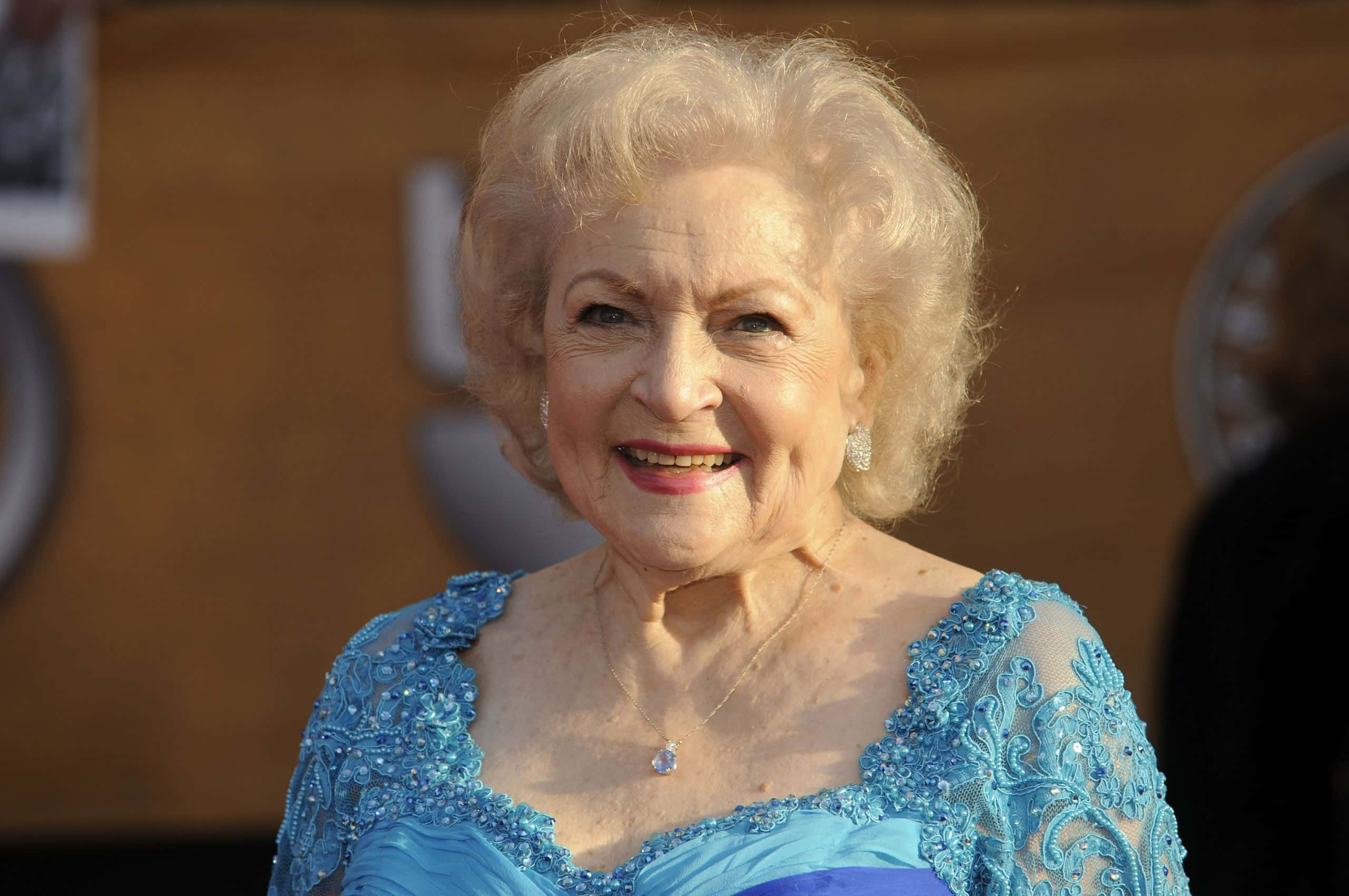 Betty White 桌布 壁紙 — 貝蒂·懷特在金色燈光下，身穿閃亮藍色禮服，在華麗活動中溫暖地閃耀。