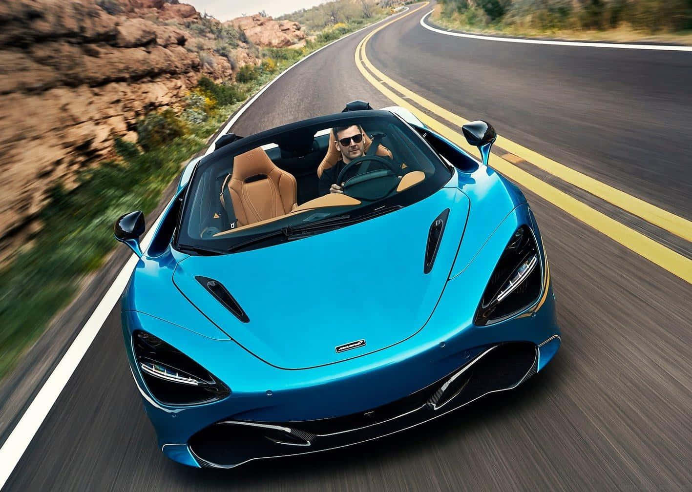 一名戴著太陽眼鏡的男子在沙漠公路上駕駛一輛藍色McLaren跑車。
