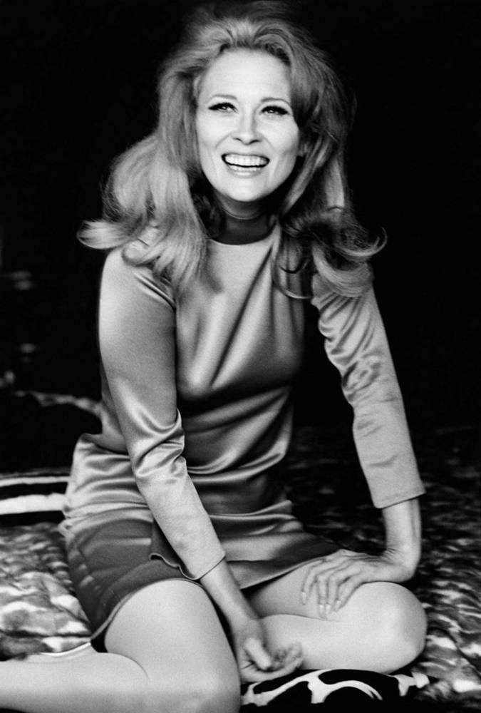 費·唐娜薇 1967 年《Vogue》雜誌封面 壁紙 — 一位容光煥發、擁有一頭豐盈金髮的女性，坐在圖案床上，在經典黑白肖像中歡快地大笑。