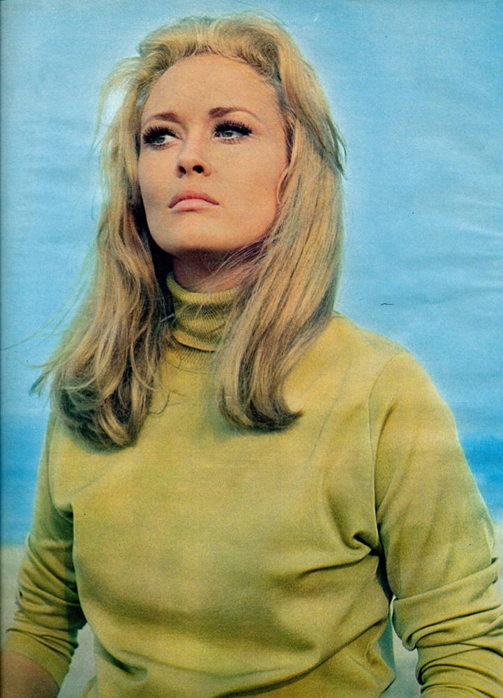 費·唐娜薇 1968 年肖像 壁紙 — 一位金髮女子穿著綠色高領毛衣，在柔和的藍天背景下向上凝視。