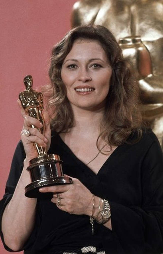 費·唐娜薇1977年奧斯卡最佳女主角獎 壁紙 — 一位微笑的黑裙女士手持金色奧斯卡獎座，背景為粉紅色，身後有一個更大的雕像。
