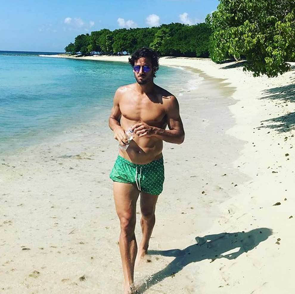 費利西安諾·洛佩斯 海灘 壁紙 — 一個肌肉發達的男人穿著綠色泳褲和太陽眼鏡，赤腳走在白色沙灘上，背景是碧綠的海水和茂密的綠樹，天空湛藍。