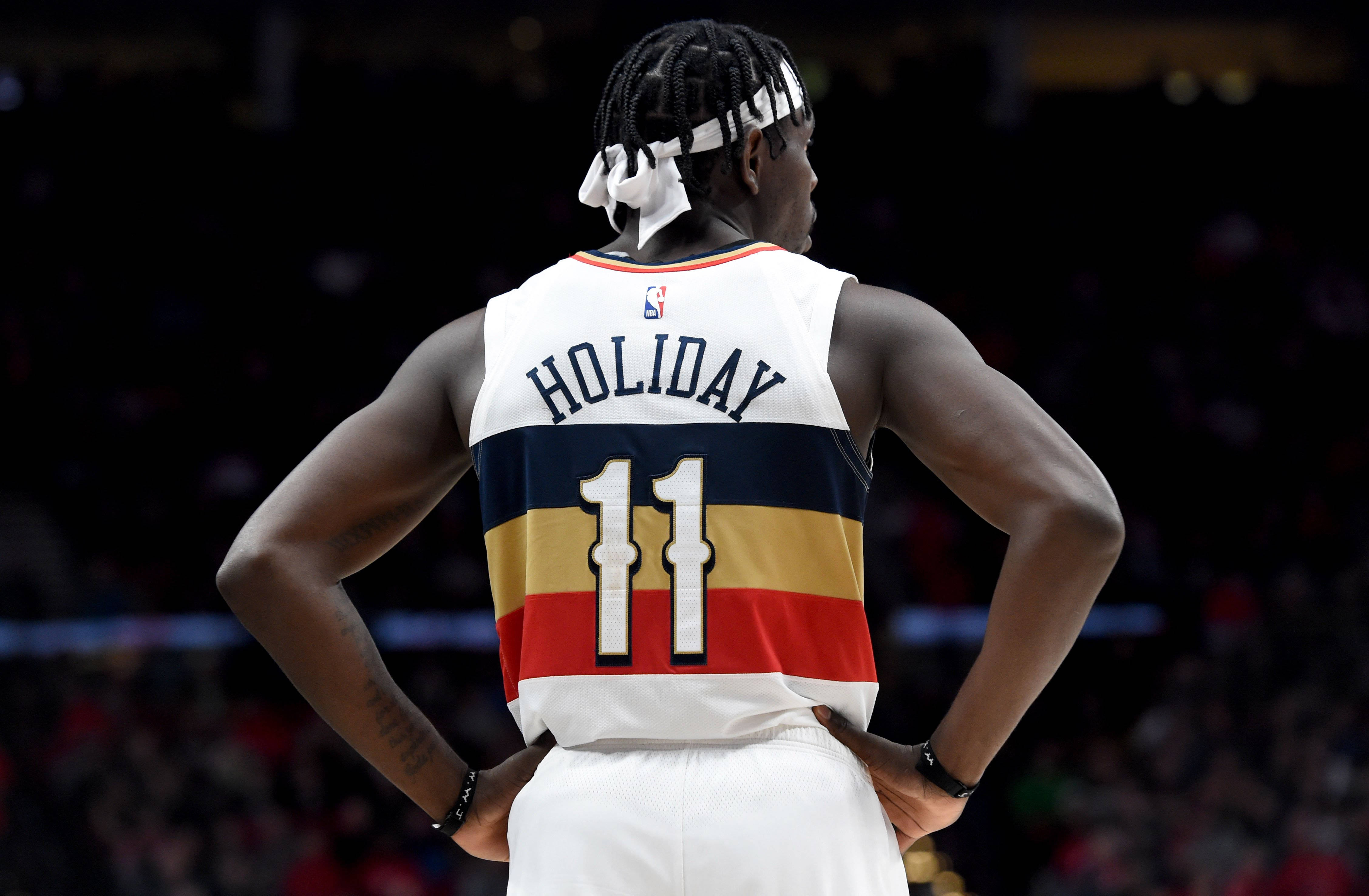 費城76人隊Jrue Holiday 壁紙 — 一名身穿白色球衣、印有「HOLIDAY 11」字樣的籃球運動員，在燈光昏暗的球場上背對鏡頭，雙手叉腰站立。