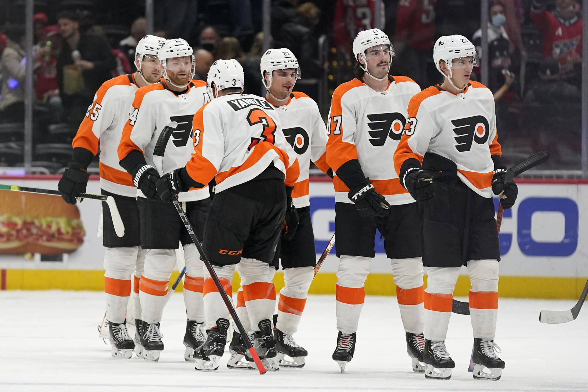 費城飛人隊 Sean Couturier 壁紙 — 身穿橙色和白色球衣的冰球運動員在比賽期間聚集在冰上，背景有球迷。
