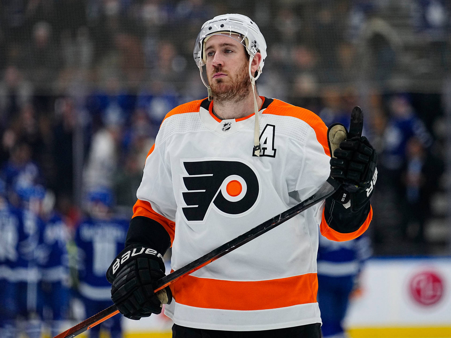 費城飛人隊凱文·海耶斯 NHL 賽事 壁紙 — 穿著白色和橙色飛人隊球衣的冰球運動員在比賽期間在冰上豎起大拇指，身後有模糊的人群。