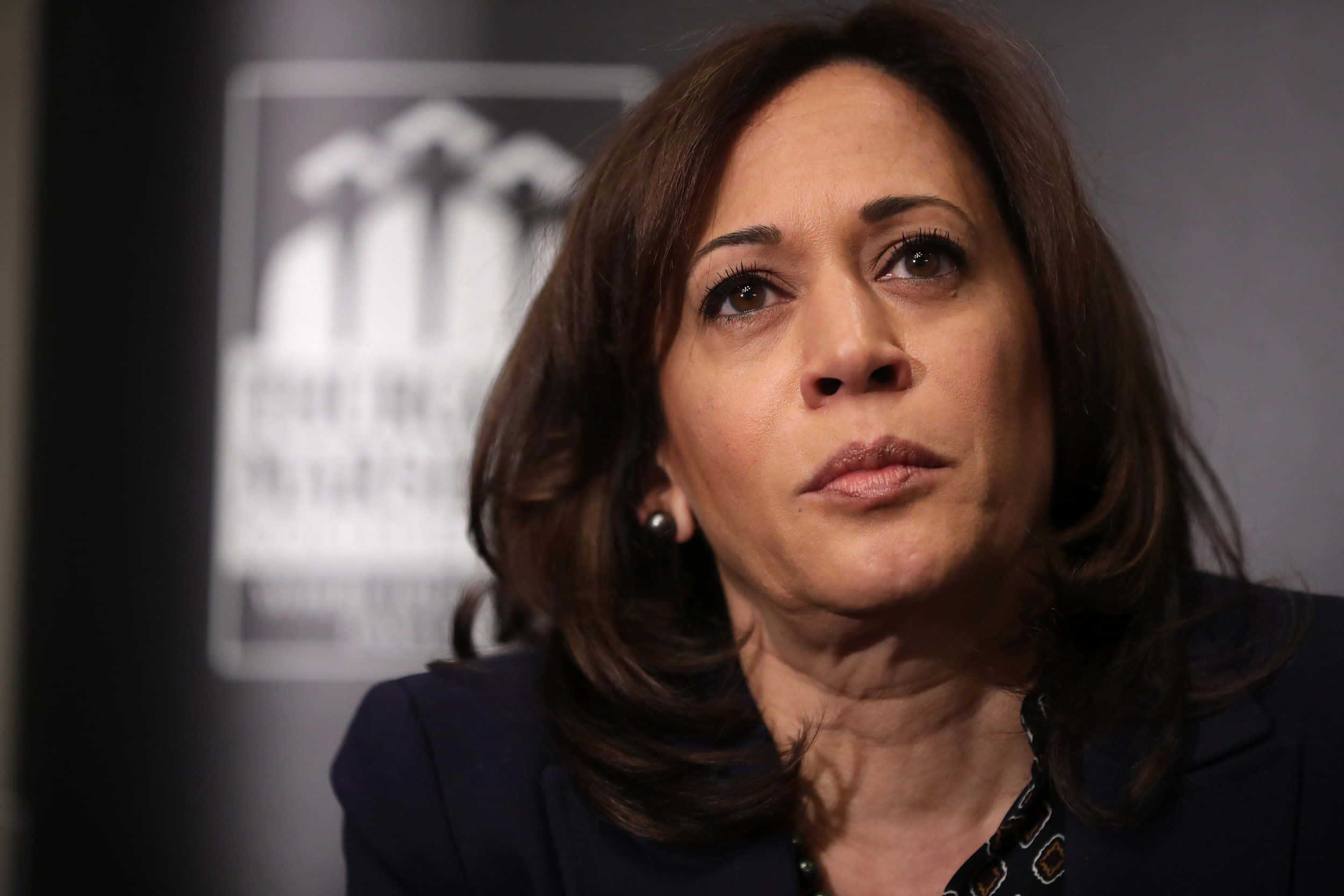 Kamala Harris 桌布 壁紙 — 賀錦麗的近距離肖像，表情嚴肅向上望，背景模糊帶有標誌。