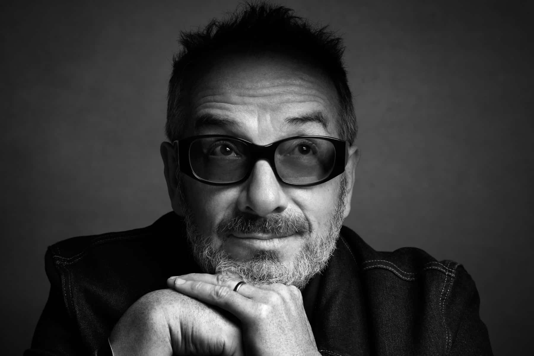 資深英國歌手Elvis Costello 壁紙 — 一個戴眼鏡的男人在純色背景下將下巴擱在手上的黑白肖像。