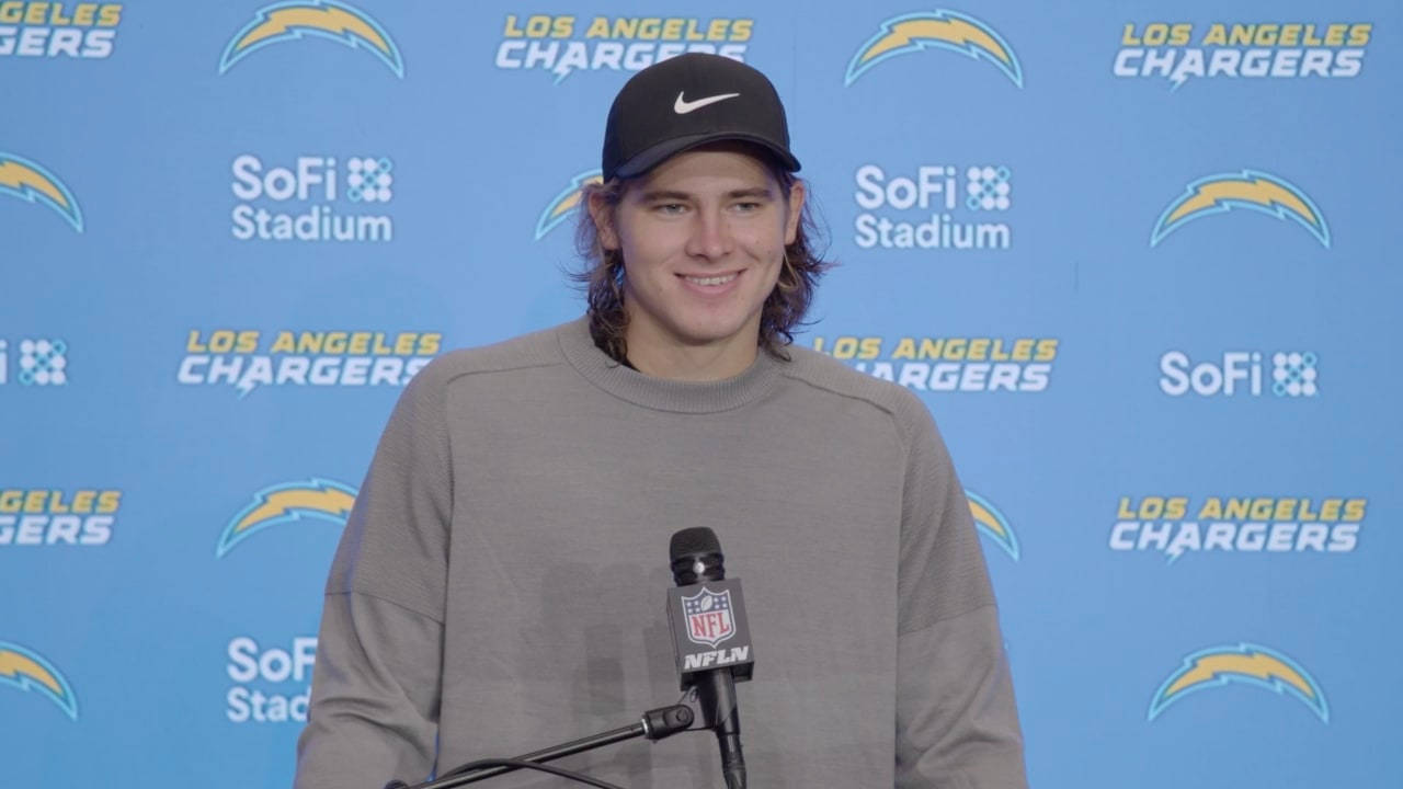賈斯汀·赫伯特 洛杉磯突襲者隊 NFL 壁紙 — 一位面帶微笑的NFL球員，戴著黑色帽子，穿著灰色運動衫，站在咪高峰前，背景是藍色的洛杉磯電光隊。