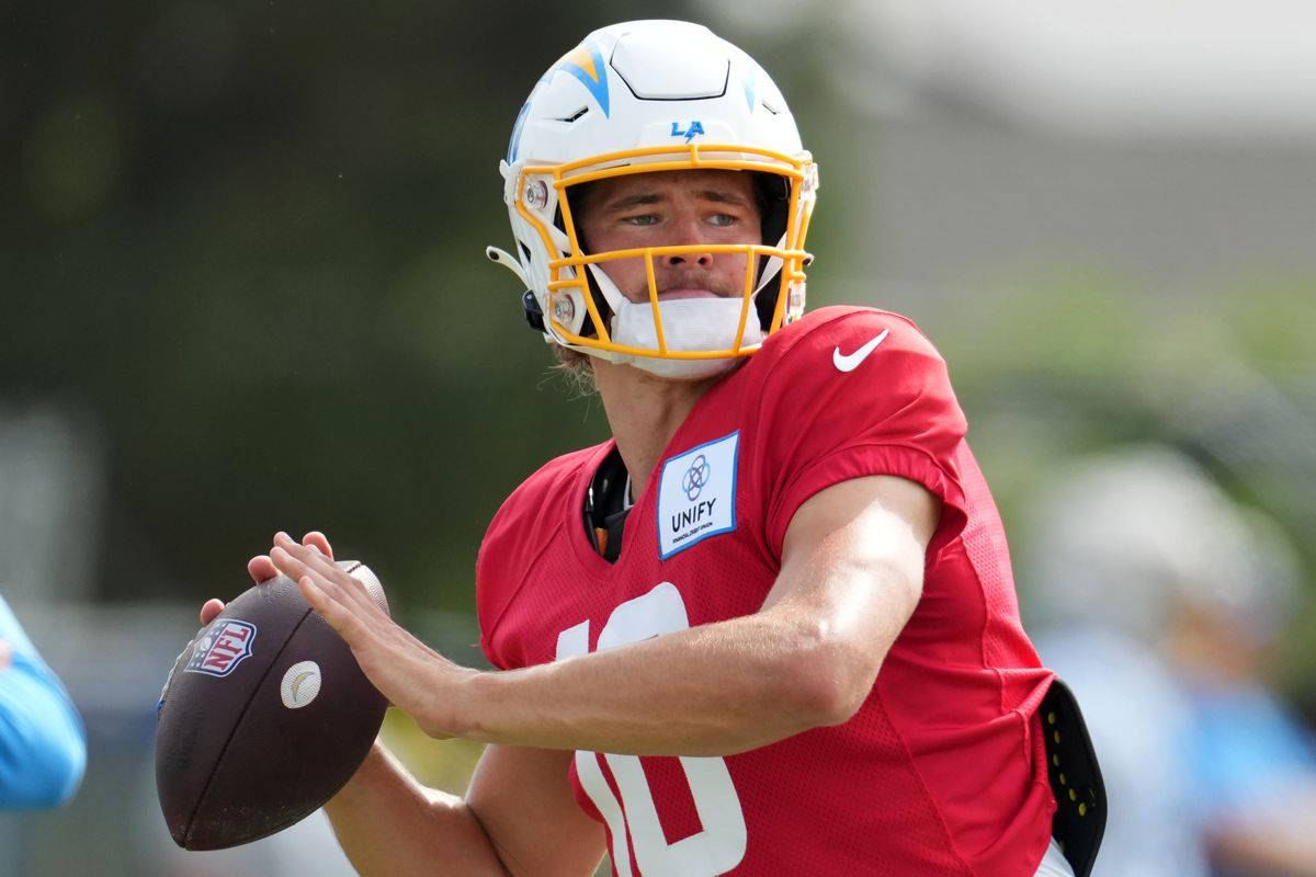 賈斯汀·赫伯特 洛杉磯突襲者隊 壁紙 — 一位專注的NFL四分衛穿著紅色球衣和白色頭盔，在陽光明媚的戶外練習中準備投擲橄欖球。