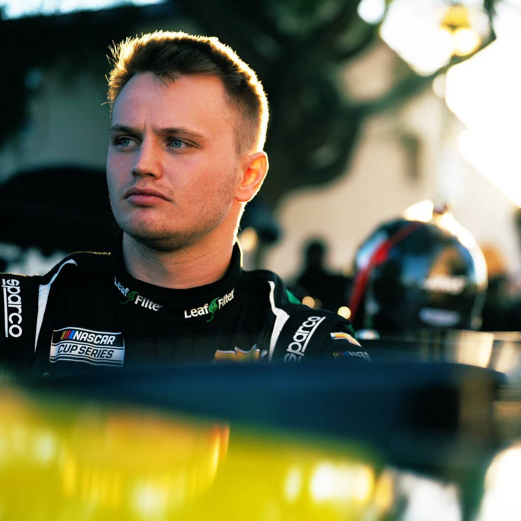 賈斯汀·黑利嚴肅神情 壁紙 — 一位專注的NASCAR車手穿著黑色賽車服，陽光照射下凸顯他的短髮和嚴肅表情，凝視著前方。