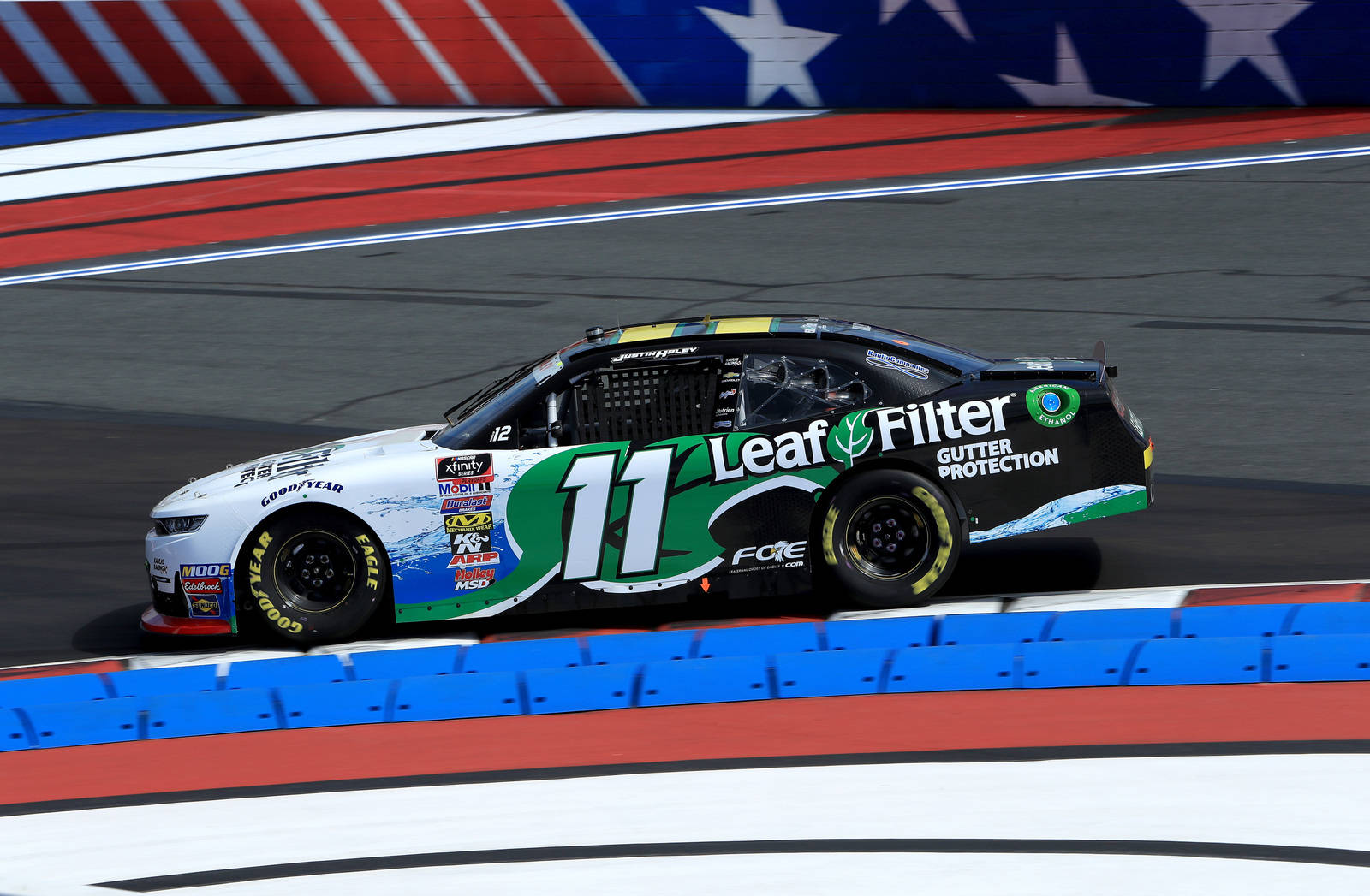 賈斯汀·黑利駕駛11號賽車高速競賽 壁紙 — 一輛帶有 Leaf Filter 品牌的 NASCAR 賽車在明亮的日光下，沿著由紅、白、藍色護欄圍繞的賽道飛馳。