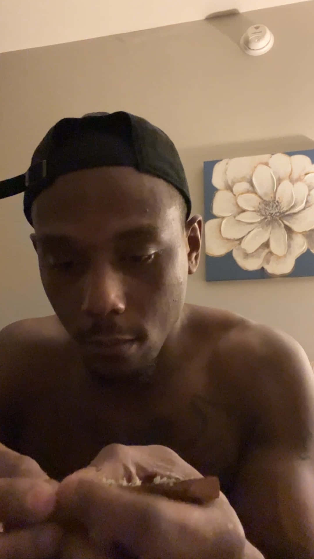 一名赤裸上身的男子戴著黑色鴨舌帽，在室內花卉畫附近拿著小雪茄。
